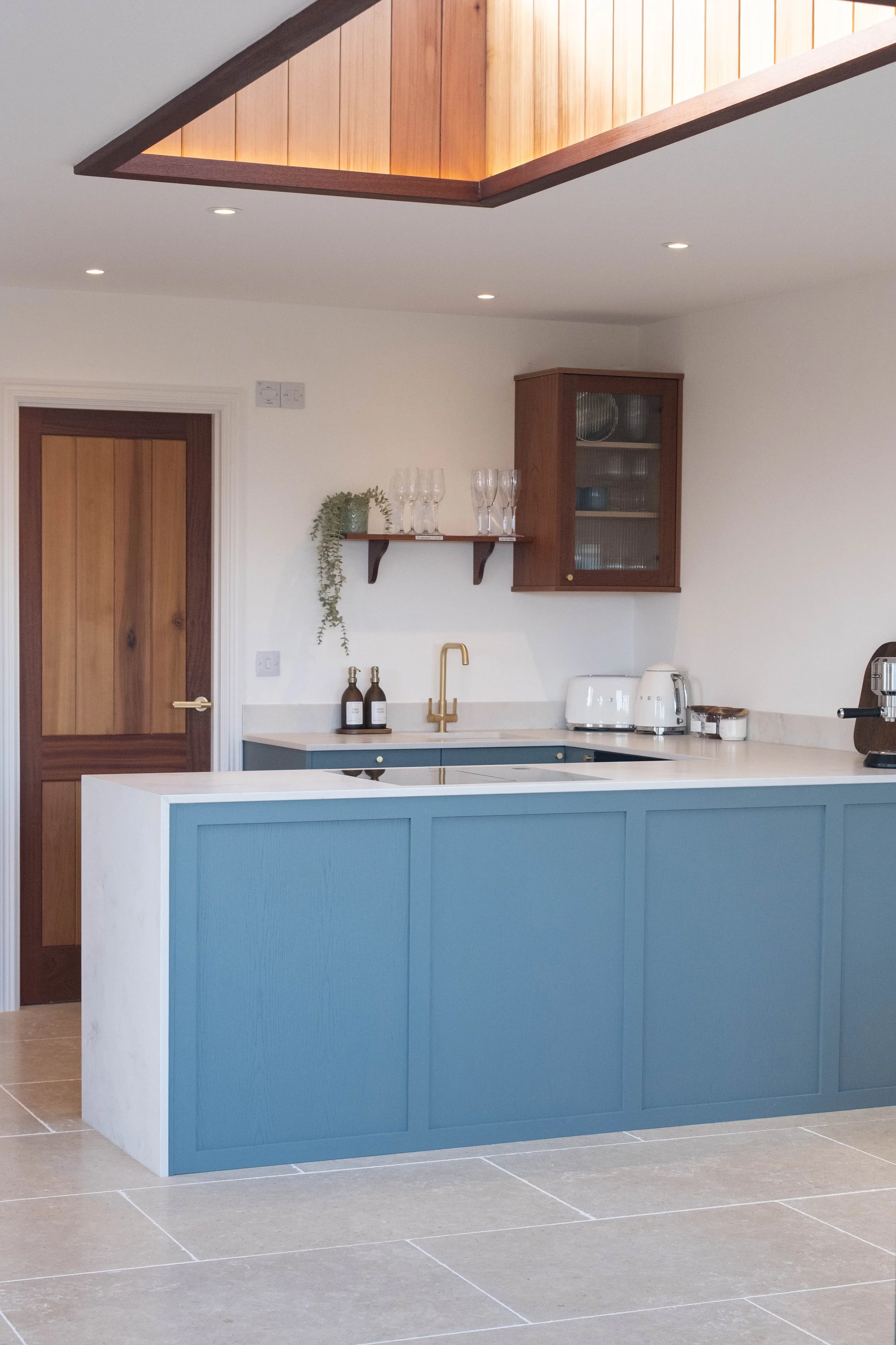 Modern kitchen with a blue and white island, hanging wine glasses, a wooden cabinet with glass doors, and a skylight with wooden panels. There are additional kitchen appliances and decorative items on the counter.