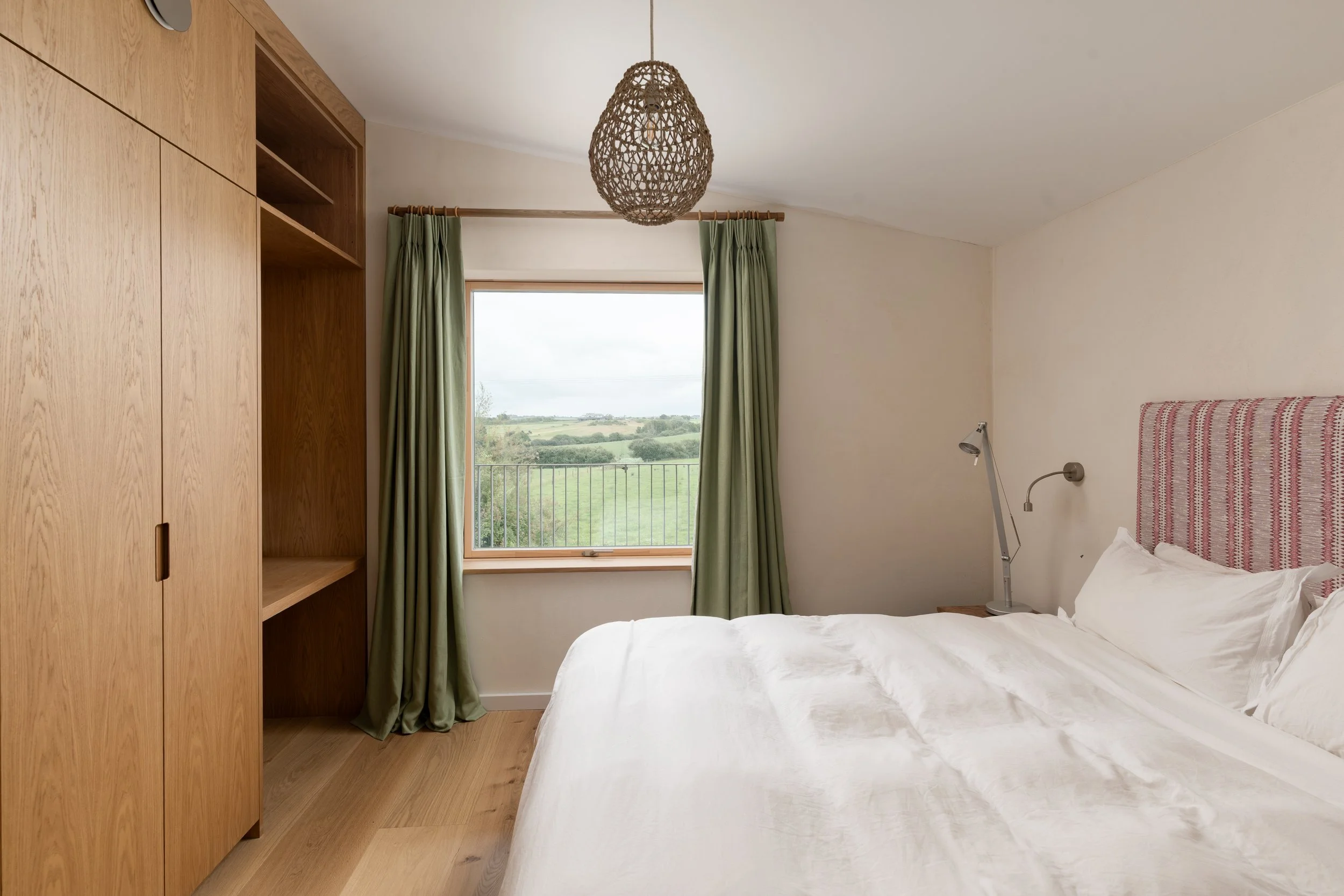 A bedroom with a large window showing green fields outside, a bed with white bedding and a striped pink and white headboard, a wooden wardrobe, and a green curtain.