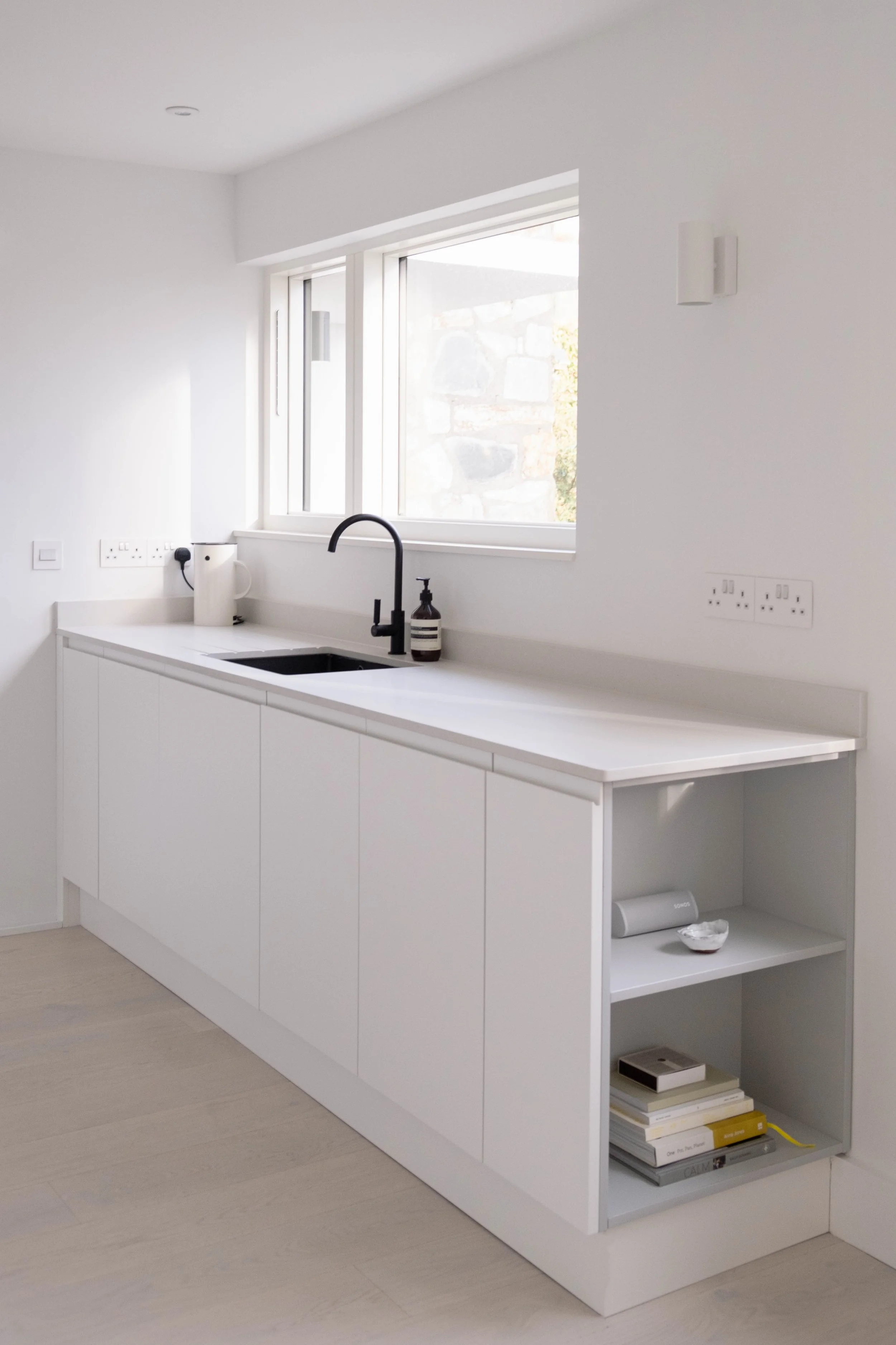 Modern white kitchen with a black faucet, white countertop, open shelving with books, a white kettle, and a window with outside stone wall and greenery.