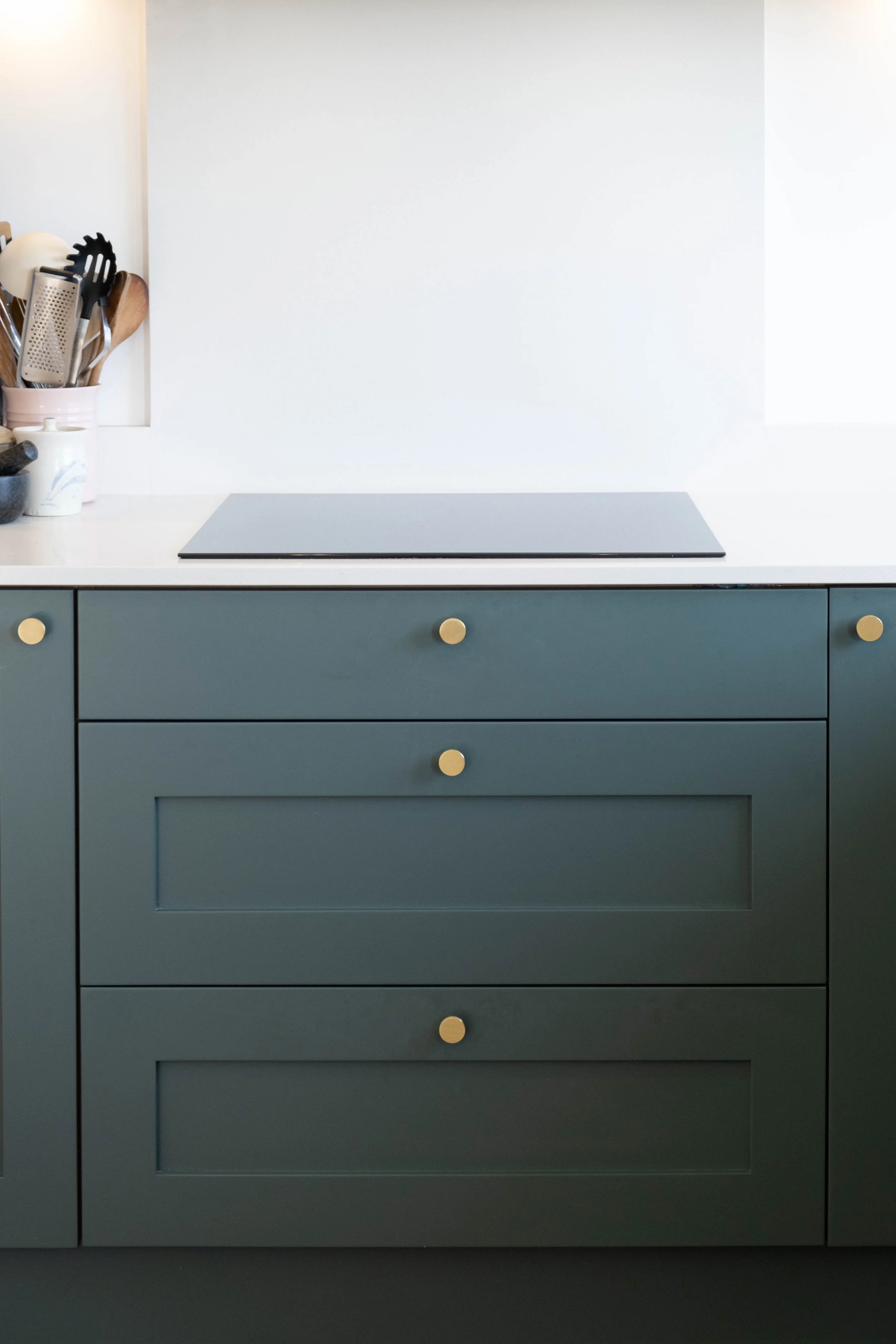 A kitchen countertop with a white surface, dark blue drawers with gold knobs, and a black electric cooktop. To the left, a white container holds various kitchen utensils, including wooden spoons, a slotted spatula, and a grater.