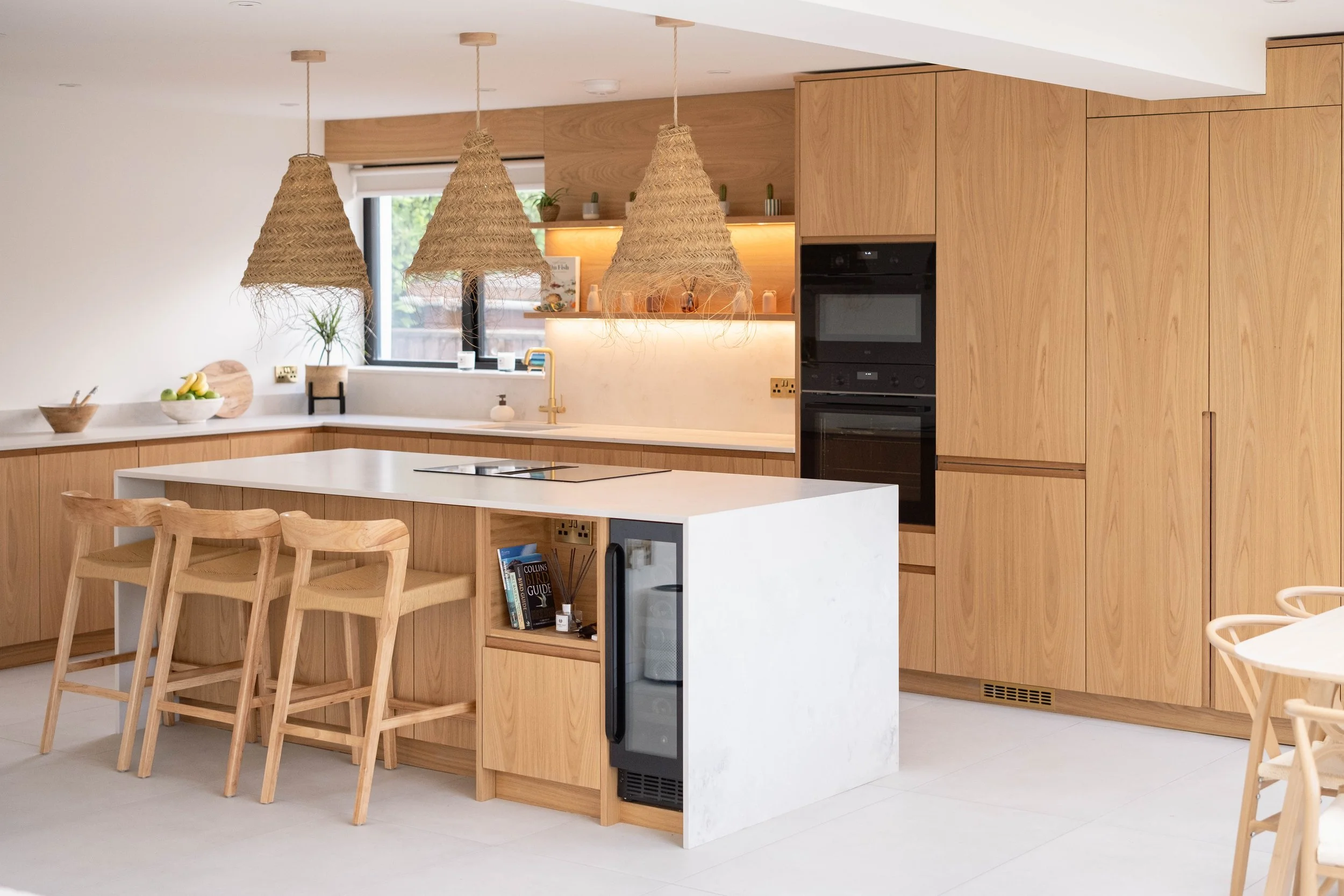 Modern kitchen with light wood cabinetry, white countertops, and three woven pendant lights hanging above an island with wooden chairs. There are plants and a window in the background.
