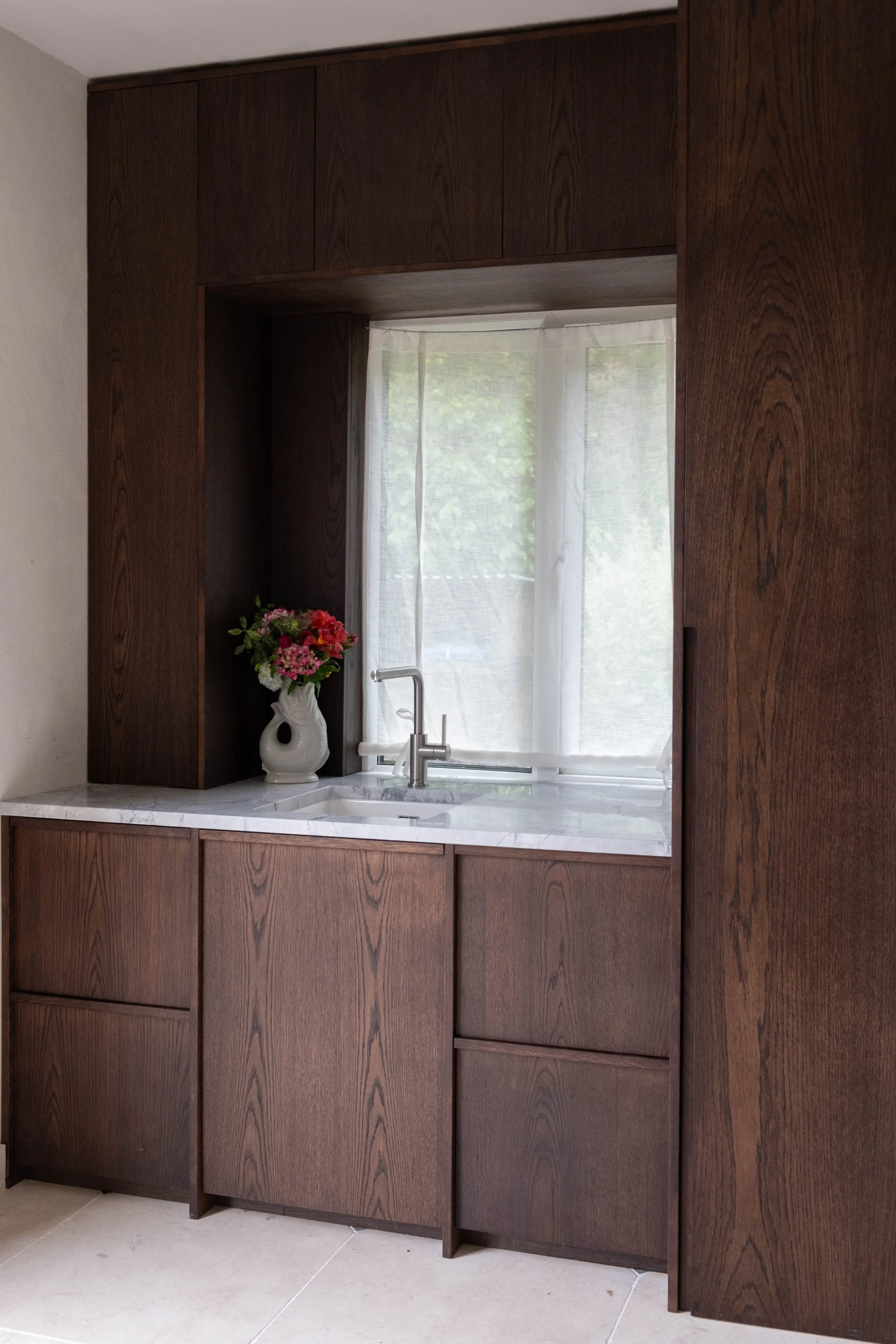 A close-up view of a kitchen window with white curtains, a marble countertop, a white ceramic vase with pink, purple, and red flowers, a silver faucet, and wooden cabinets with a dark brown finish.