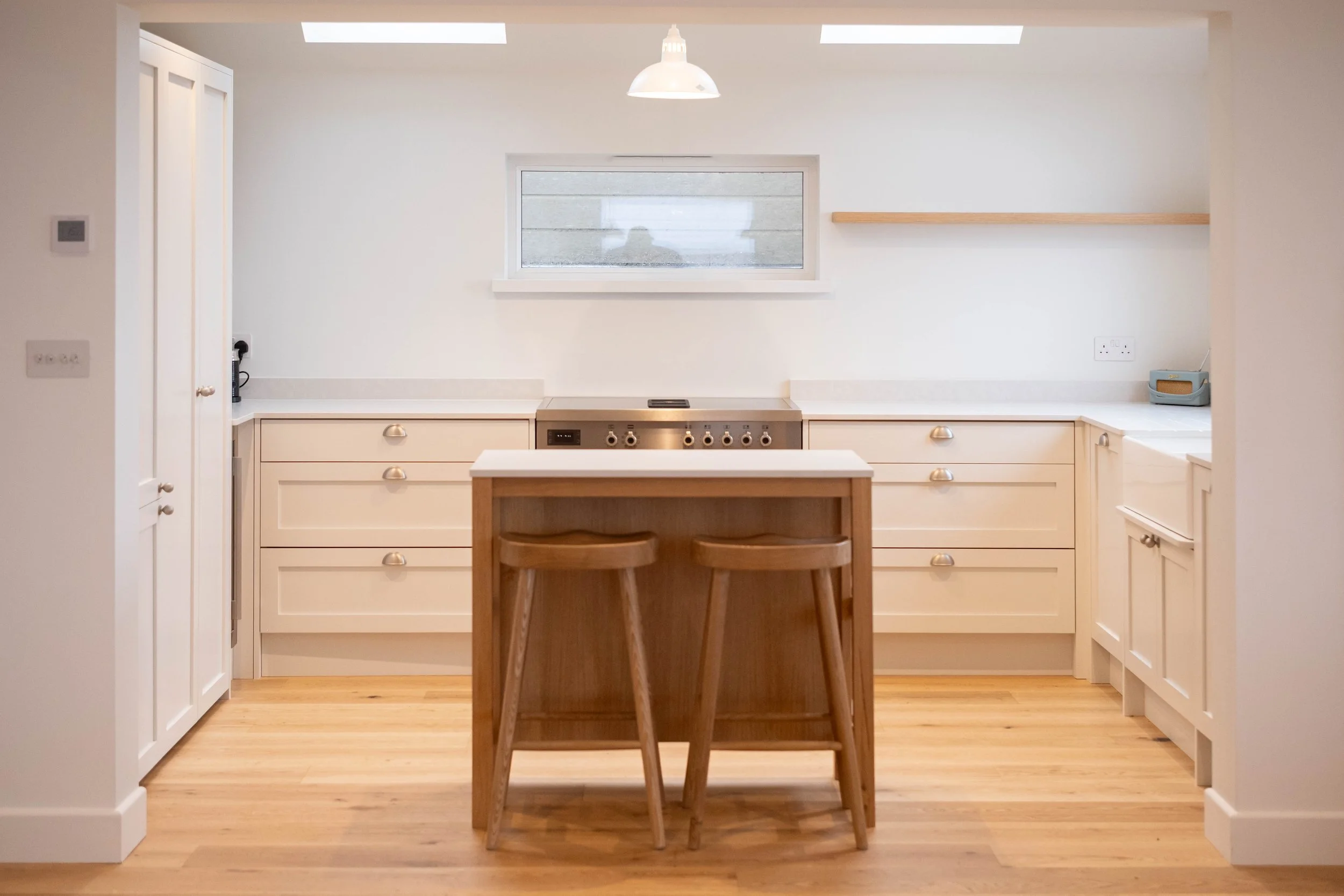 A minimalist kitchen with white cabinets, a stove, a window, and a small kitchen island with two wooden stools.