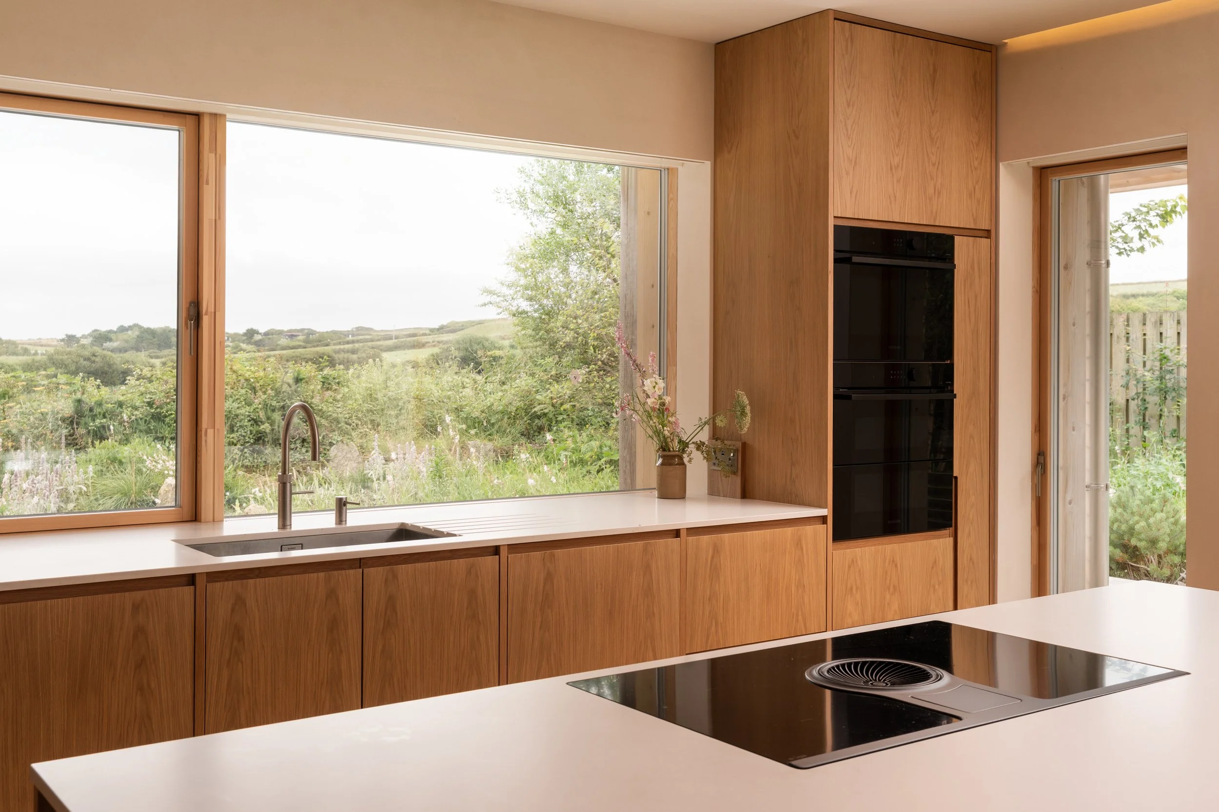 A modern kitchen with large windows overlooking a lush green landscape, featuring wooden cabinets, a white countertop, a stainless steel sink, and a black stovetop.