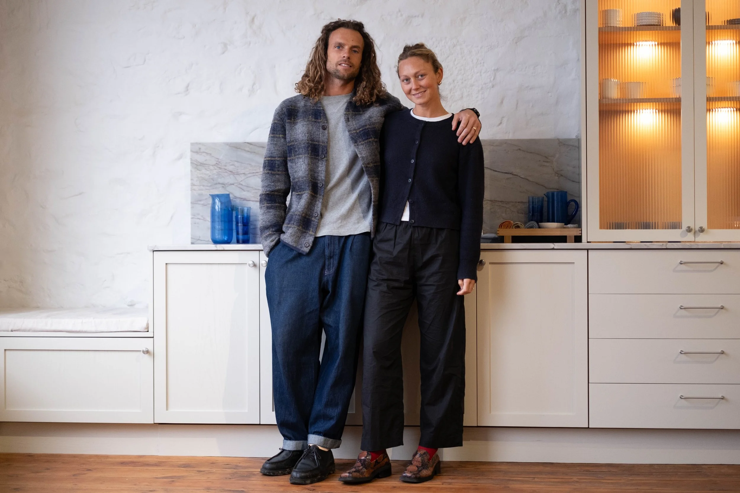 A man and woman stand close together in a kitchen, smiling at the camera. The man has long curly hair, a beard, and is wearing a gray shirt with a plaid jacket and blue jeans. The woman has her hair tied back, and is wearing a black cardigan, white shirt, and plaid shoes.