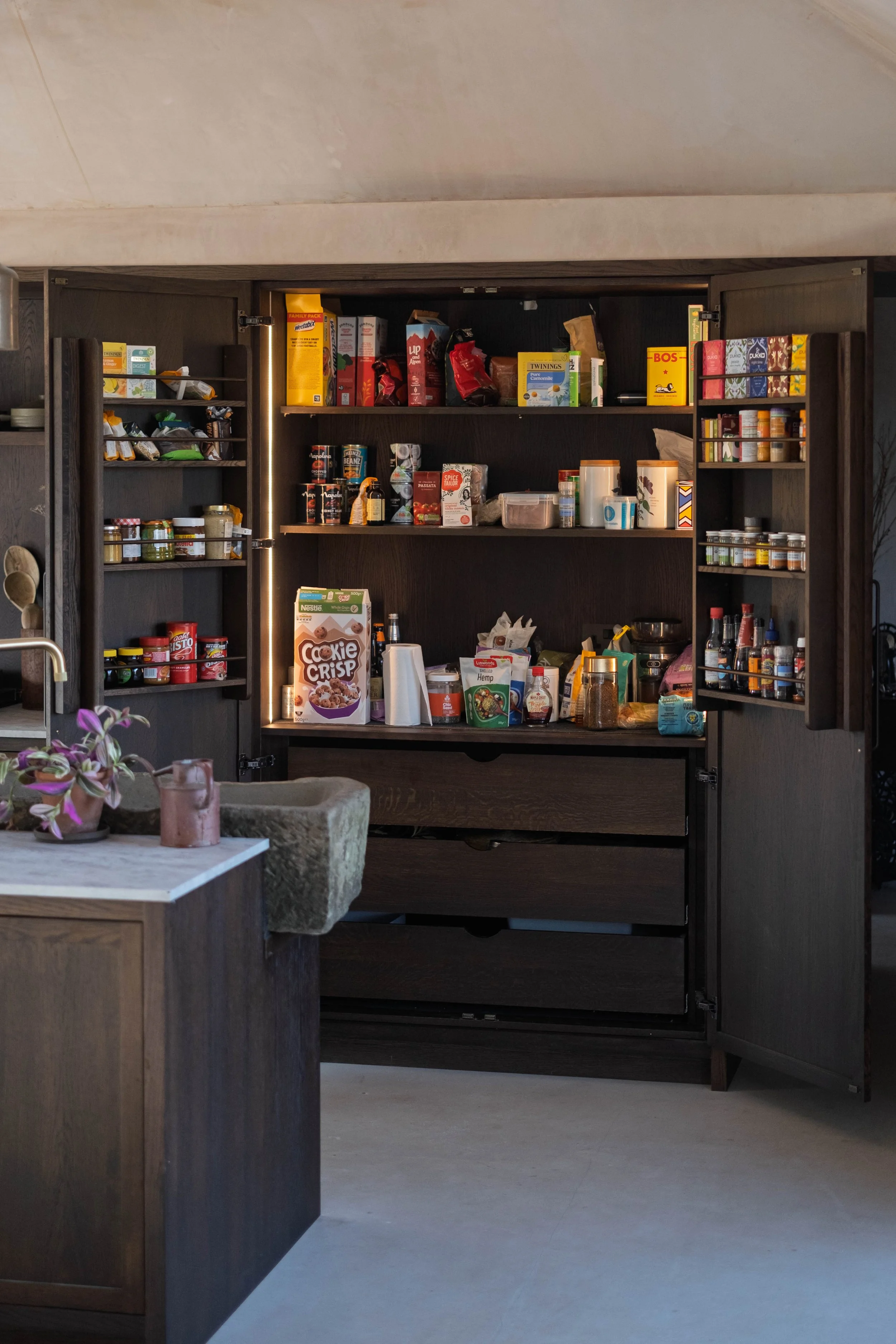 Open kitchen cabinet filled with various pantry items, spices, jars, and boxes, with some items on the kitchen counter below.