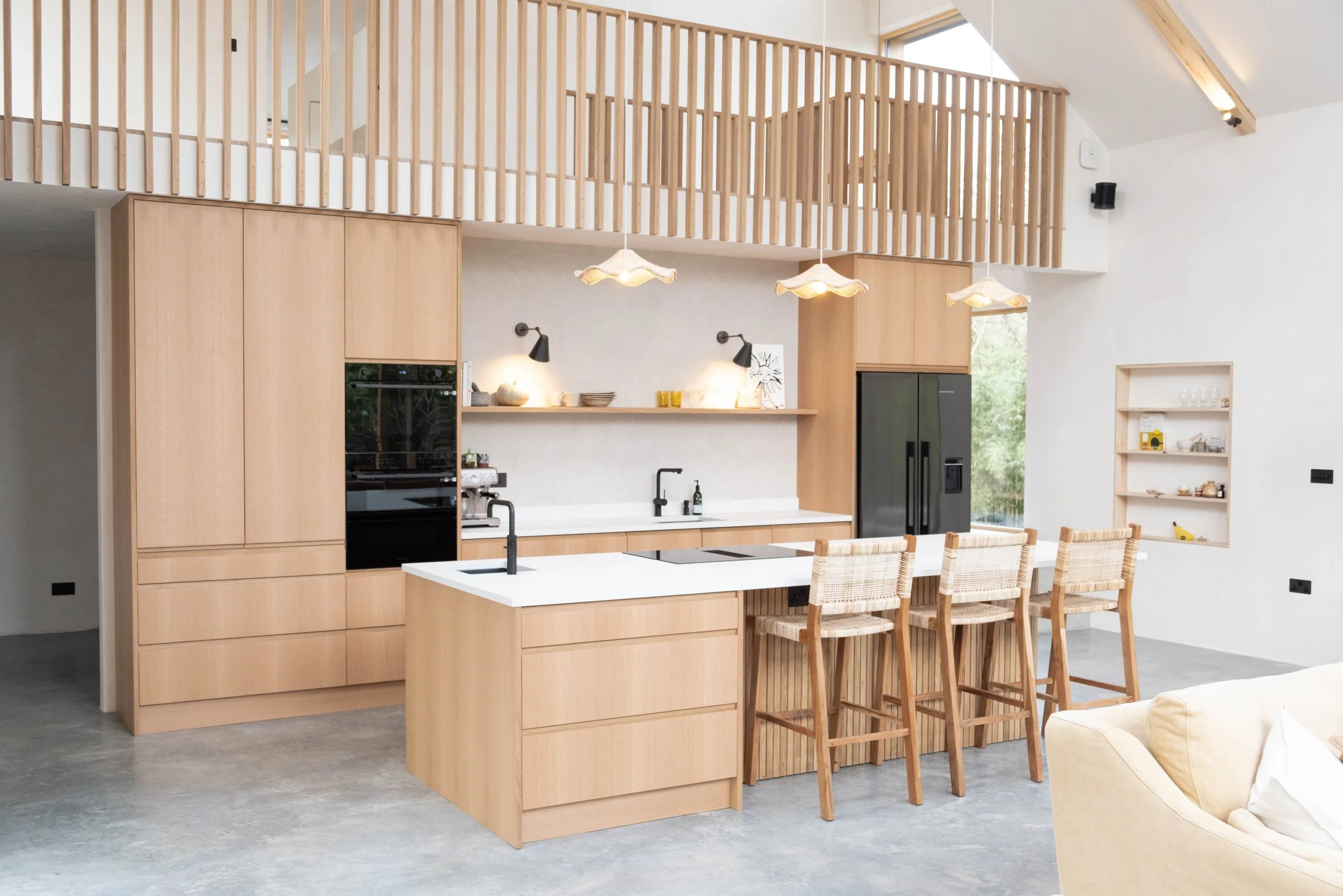 Open-plan modern kitchen with wooden cabinetry, a kitchen island with a white countertop, dining chairs, and pendant lighting with fabric shades. There is a black oven, a black refrigerator, and built-in shelves on the right side. The space features 