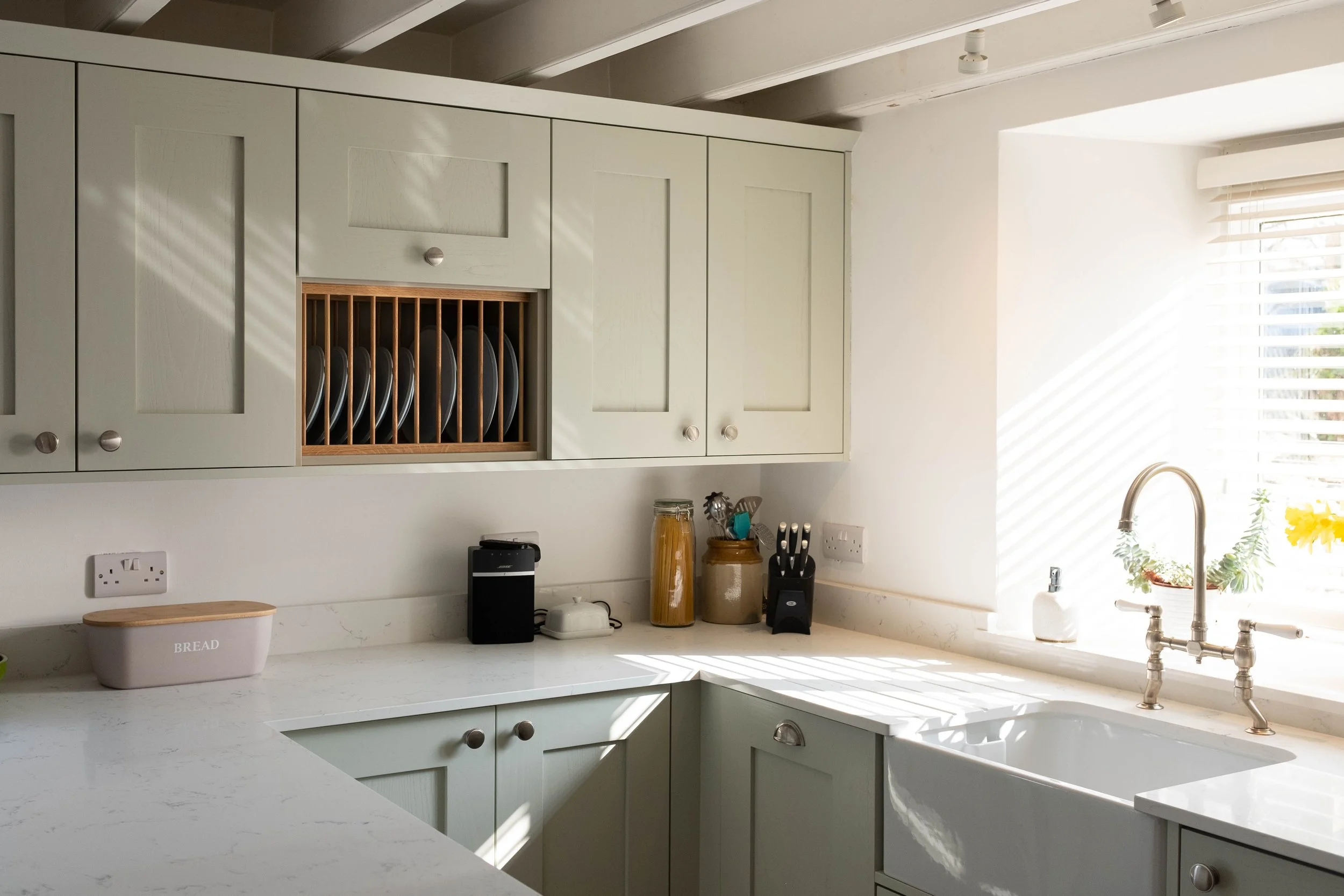 Kitchen with white cabinets, a white countertop, a dish rack with plates, and a window with sunlight streaming through blinds.
