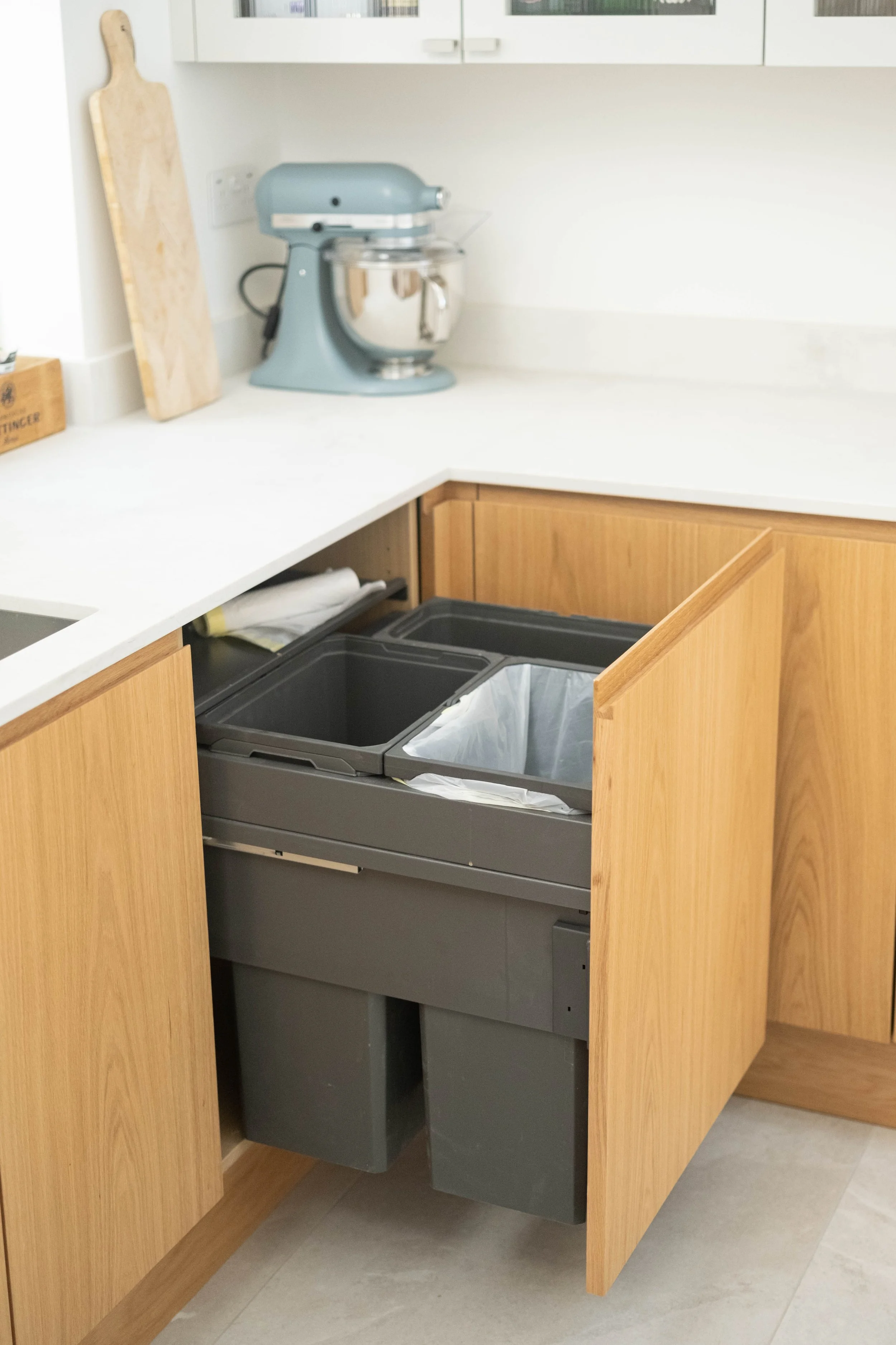 Kitchen cabinet with two open doors revealing trash and recycling bins inside, with a stand mixer and cutting boards on the countertop.