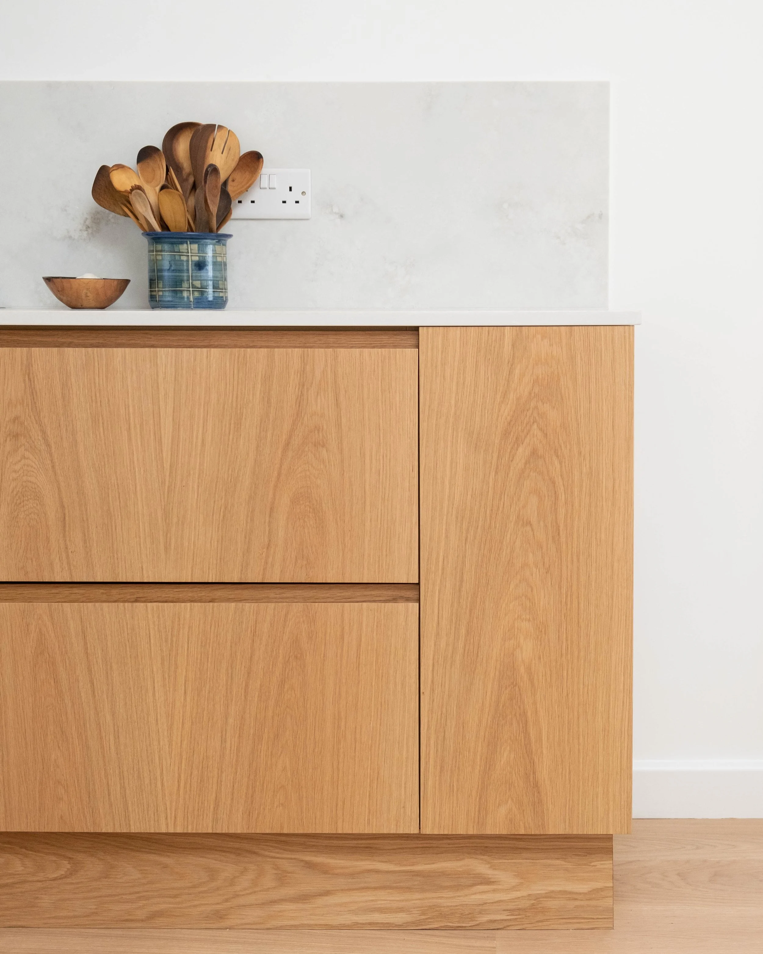 Wooden kitchen cabinet with a small ceramic bowl and a jar filled with wooden cooking spoons, near an electrical outlet on the wall.
