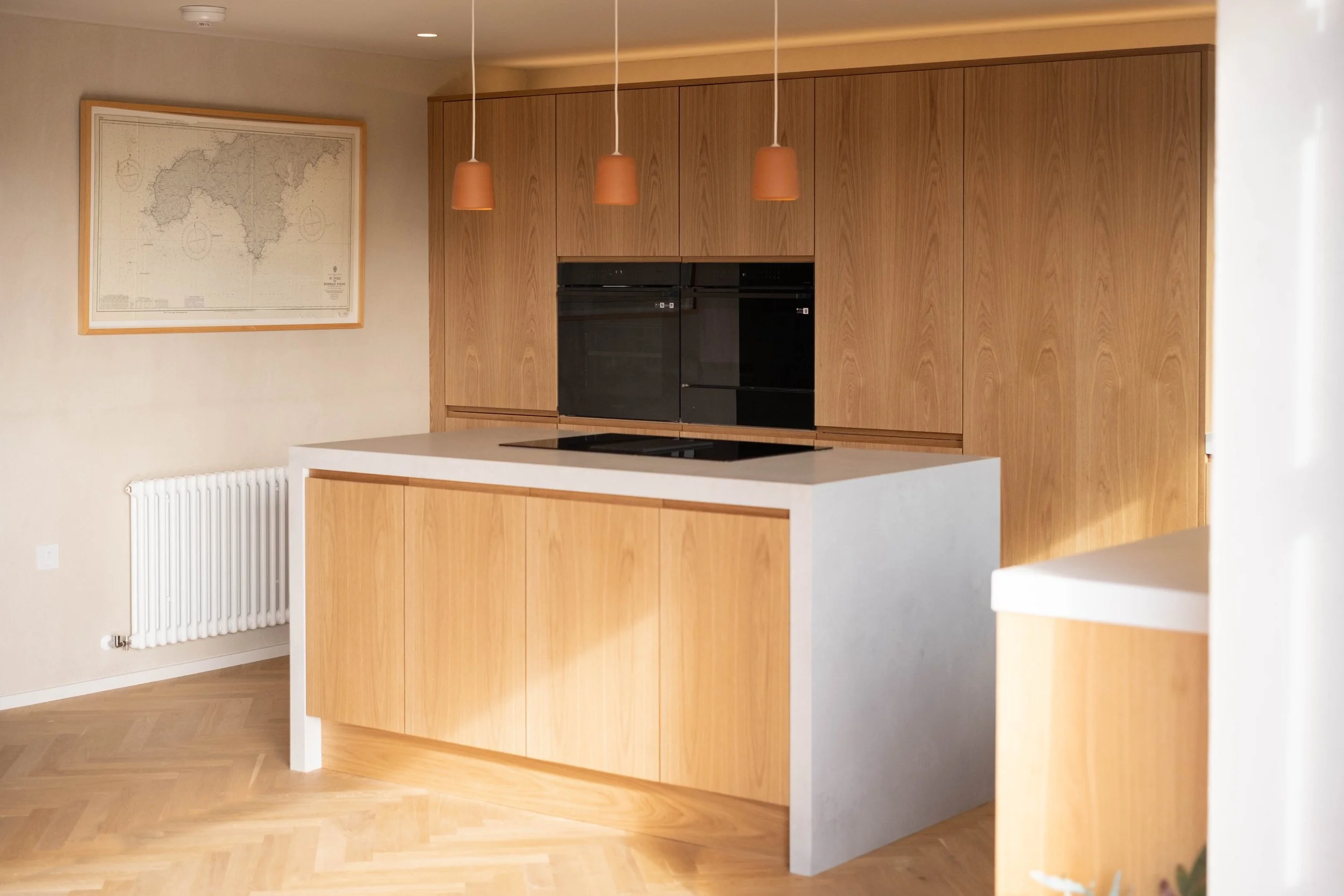 Modern kitchen with wooden cabinets, a white countertop island, three hanging pendant lights, a wall map, and a white radiator.