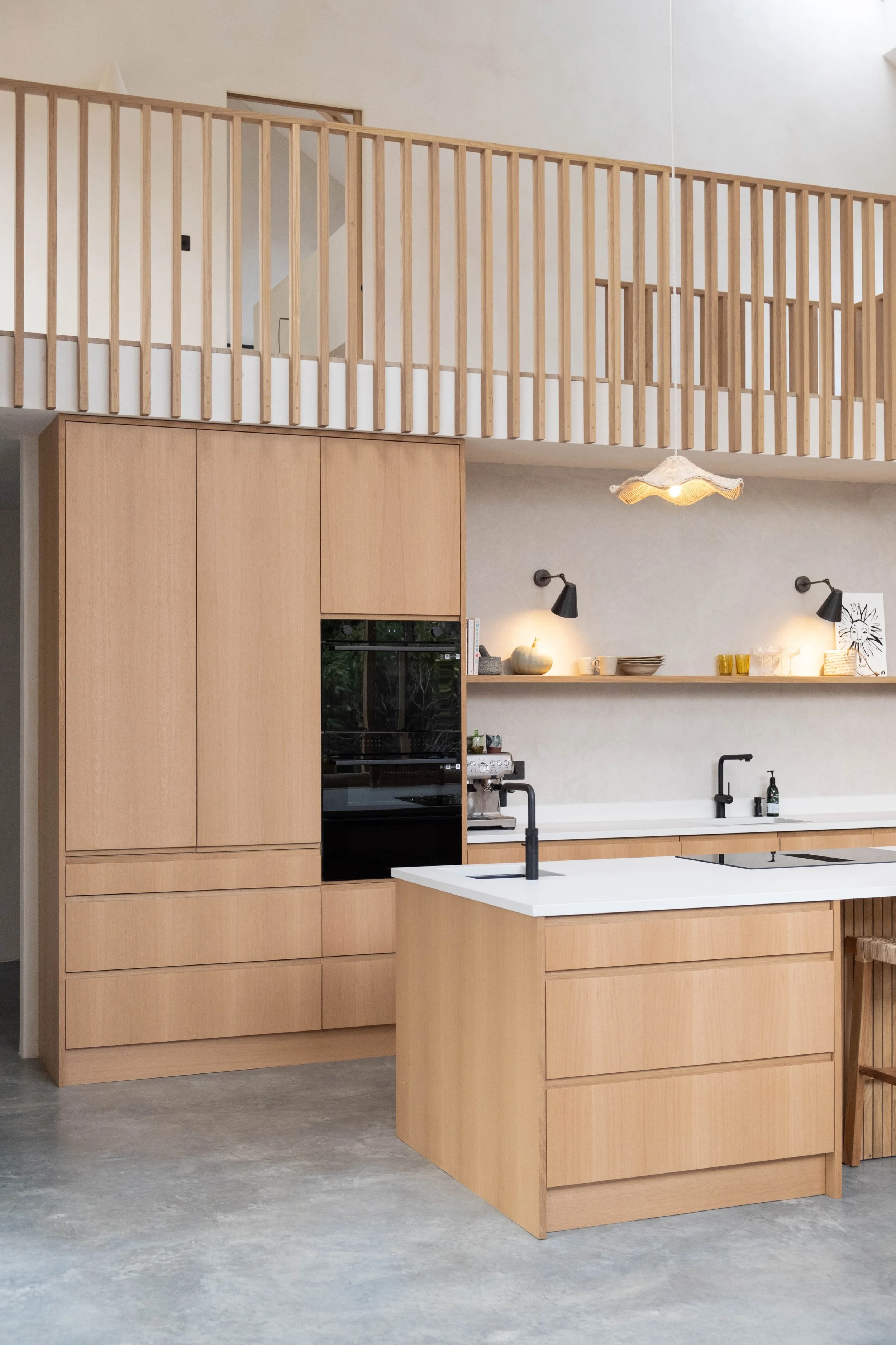 Modern kitchen with light wood cabinetry, white countertop island, black faucet, black wall sconces, and open wall shelves with decorative items. Upper level has a wooden railing, and there is a hanging pendant light.