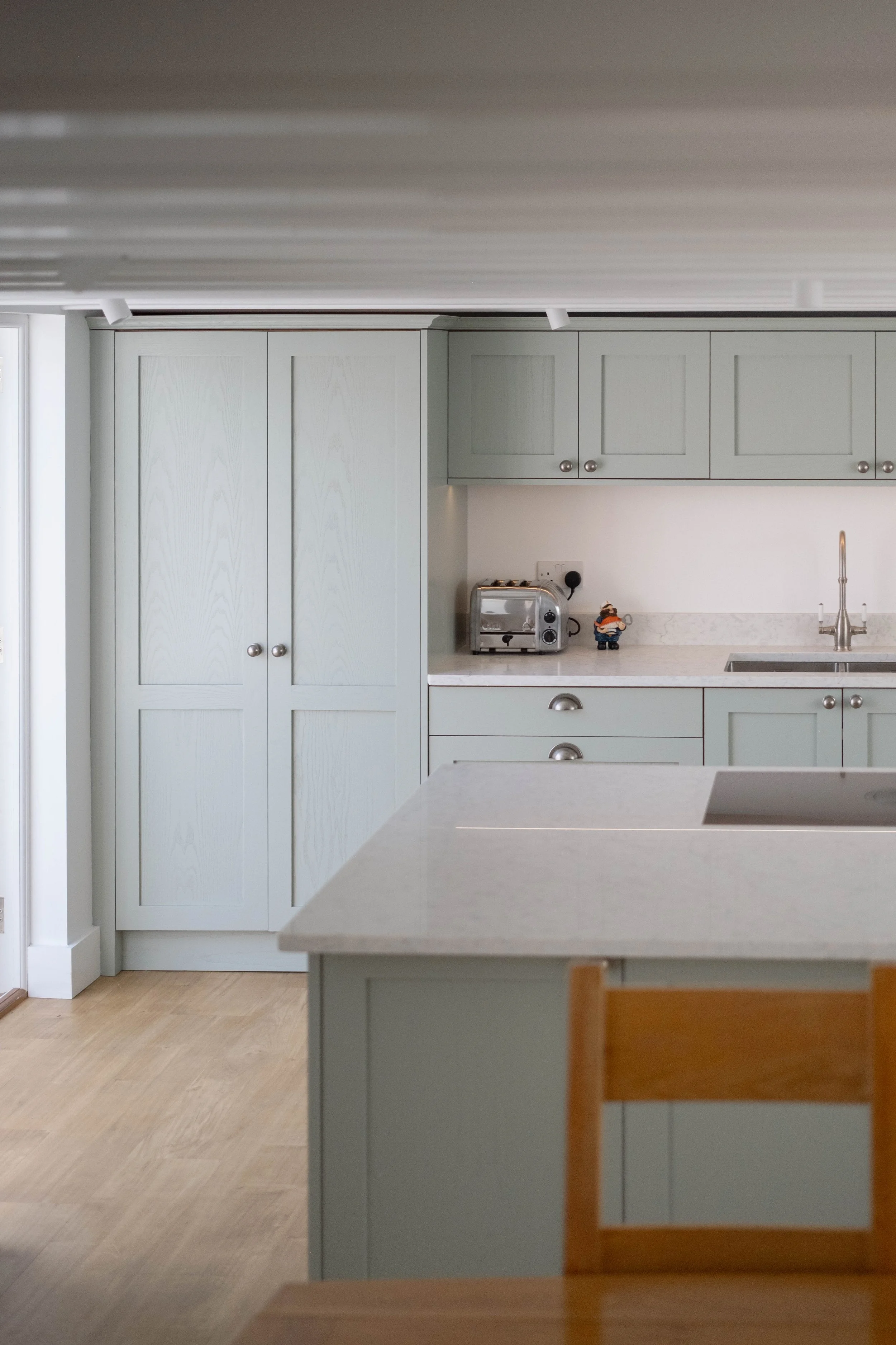 A modern, light gray kitchen with closed cabinet doors, a countertop with small appliances, and a wooden chair.