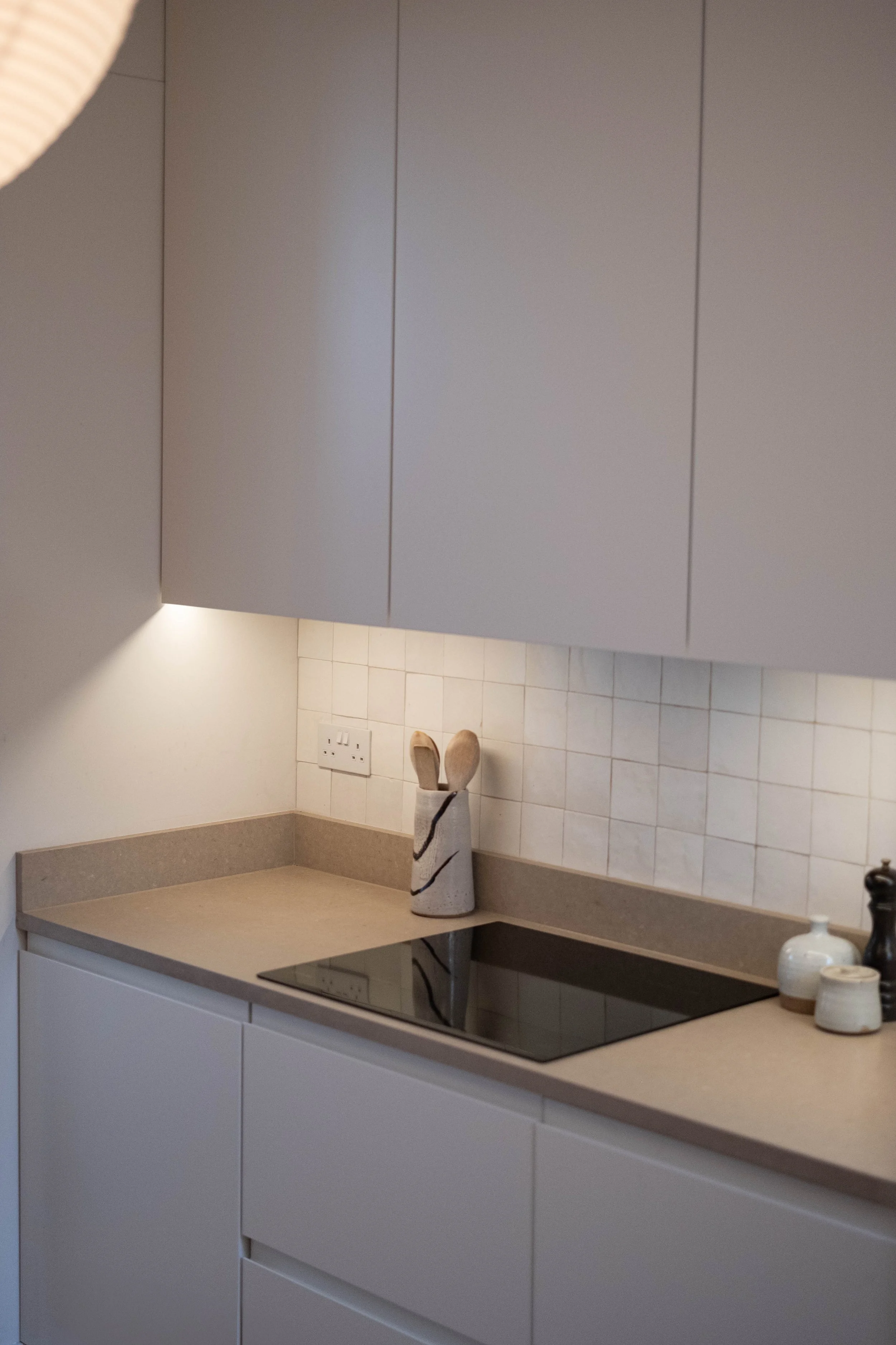 Kitchen with white cabinetry, beige countertop, and a white vase with a black stripe pattern on the counter. There are two bottles, possibly soap and lotion, on the right side of the counter. The wall has white tiles with beige grout, and an electric