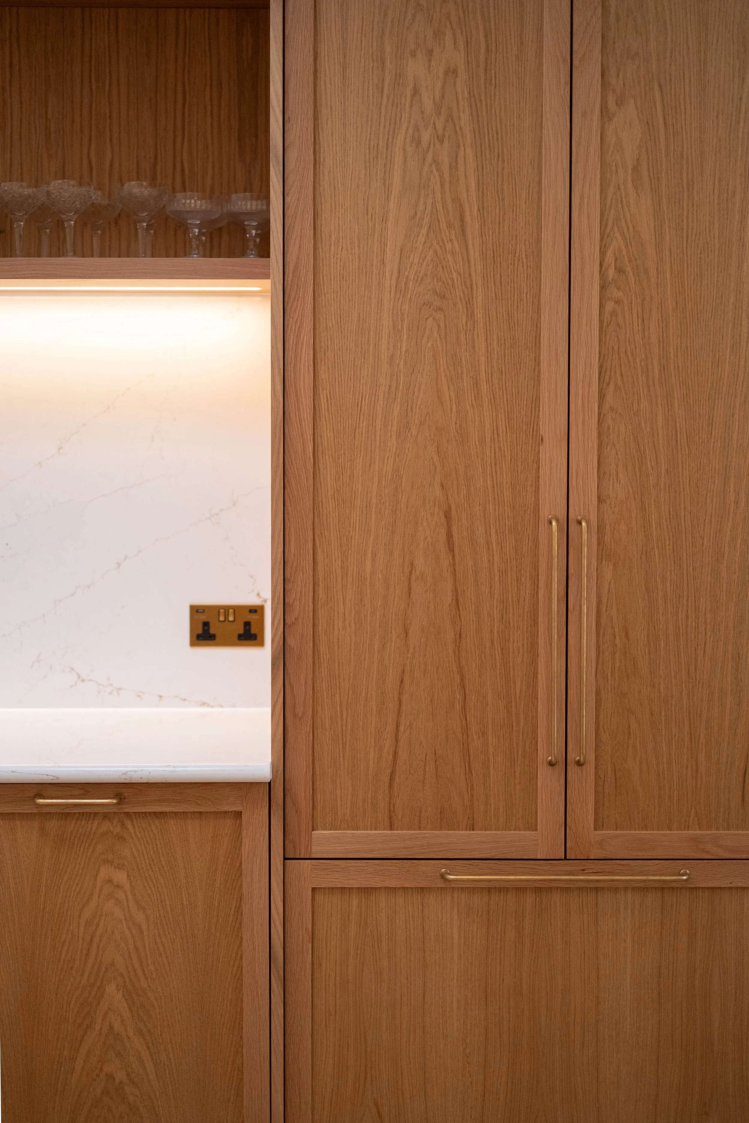 Kitchen cabinetry with wooden doors and handles, a white marble countertop, a two-plug electrical outlet, and glassware on an upper shelf.
