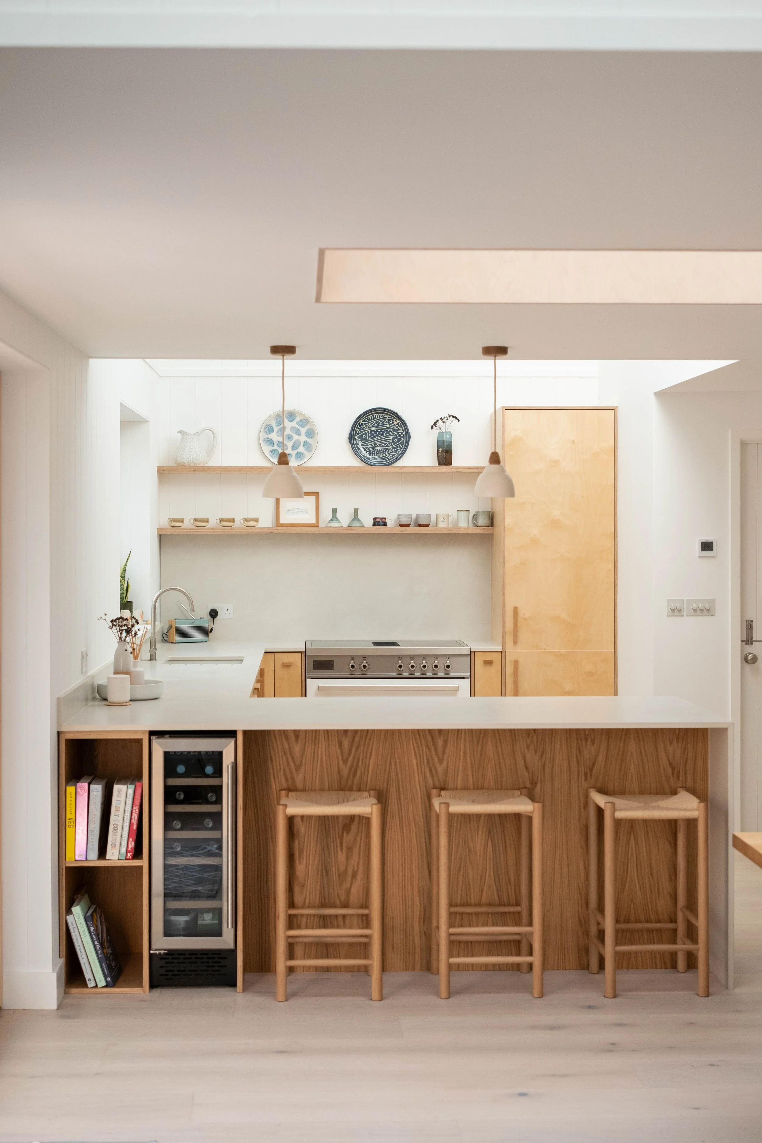 A modern kitchen with a white countertop, wooden bar stools, and a built-in wine cooler. There are open shelves with decorative plates, vases, and cups. Kitchen appliances include a stovetop, oven, and microwave. The space has minimalist design with 