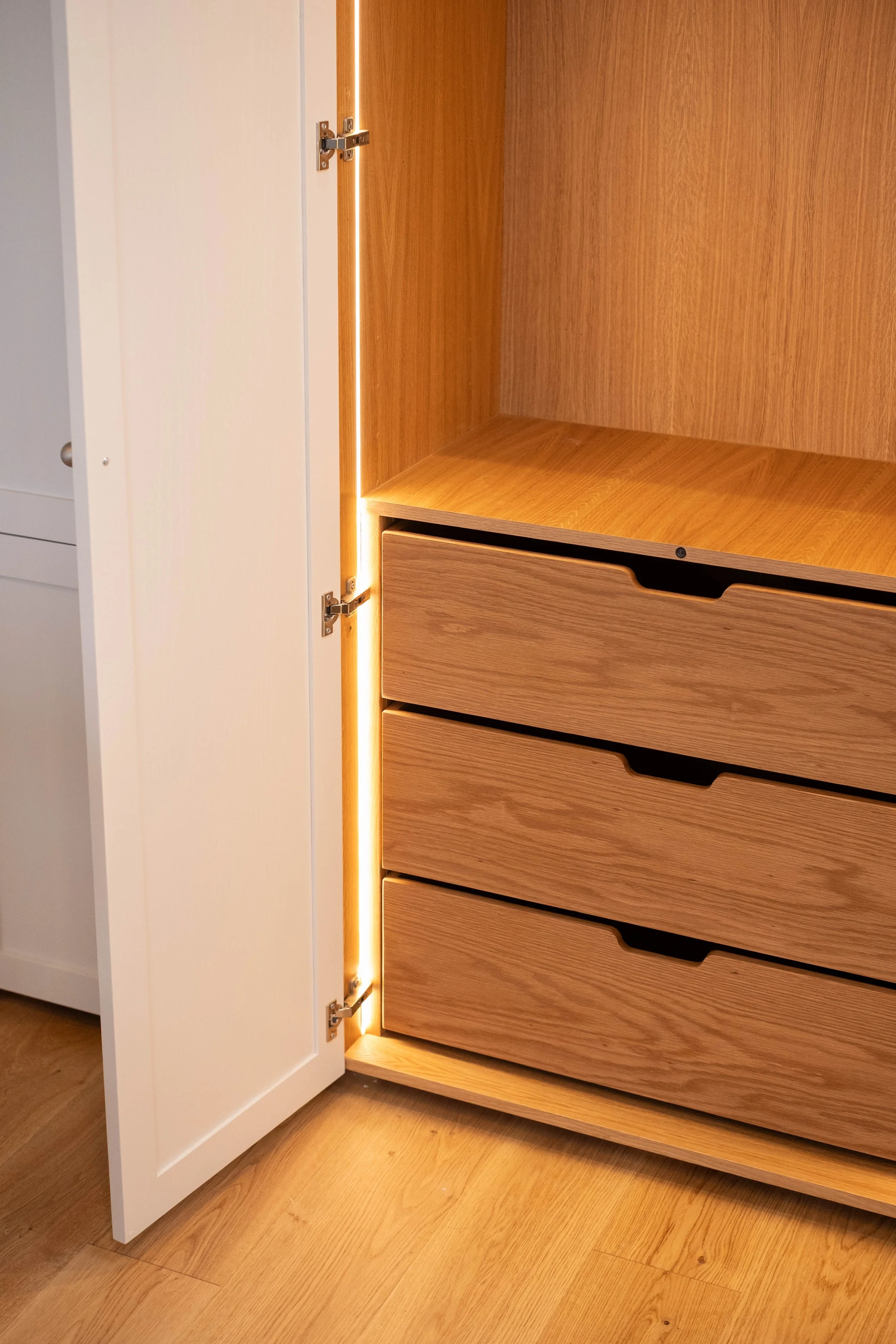 Close-up of a light wood dresser with open drawers and LED lighting along the side, next to a white door with hinges.