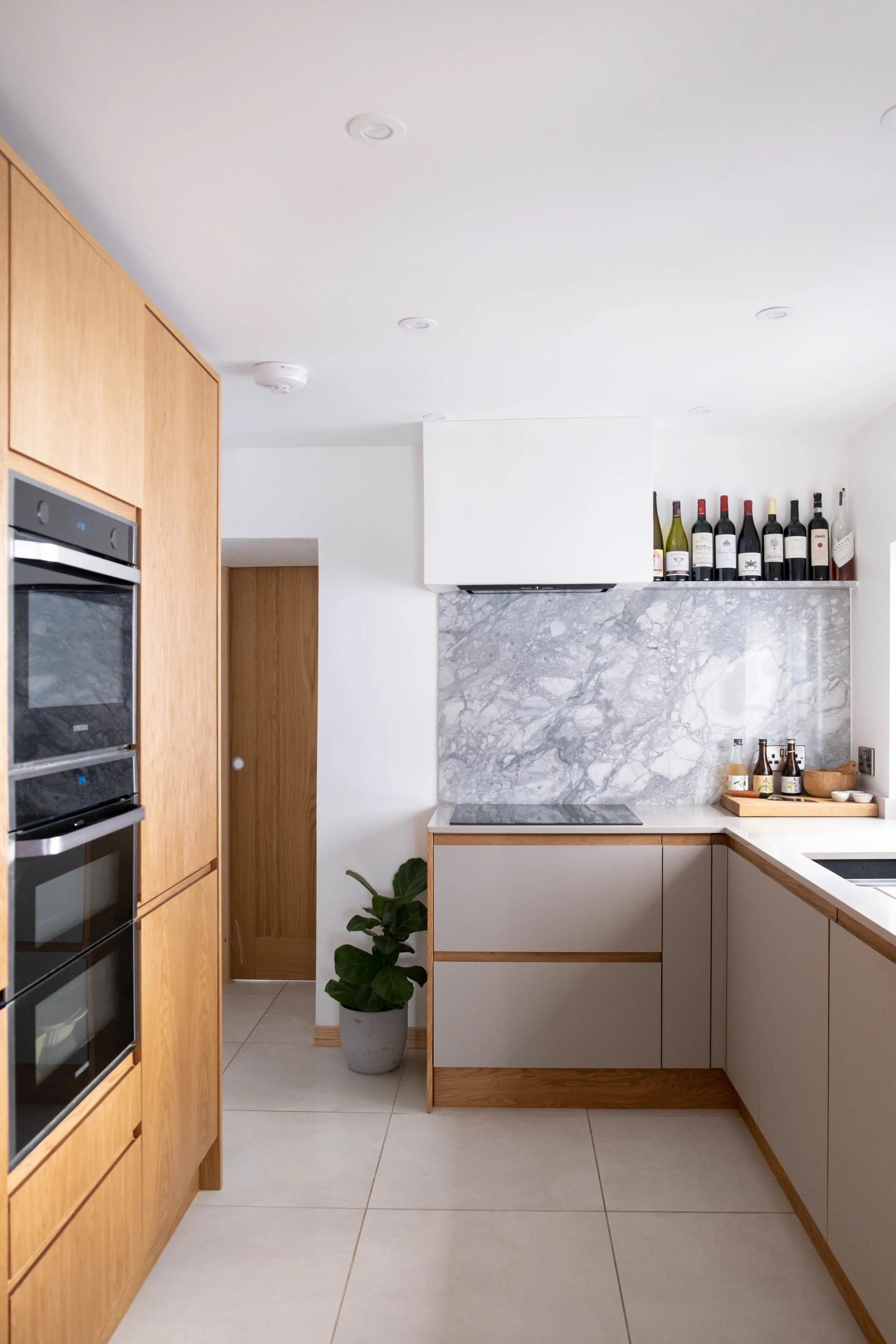 A modern kitchen with white cabinets, a marble backsplash, a row of wine bottles on a shelf, a green potted plant, and a wooden countertop with bottles and bowls.