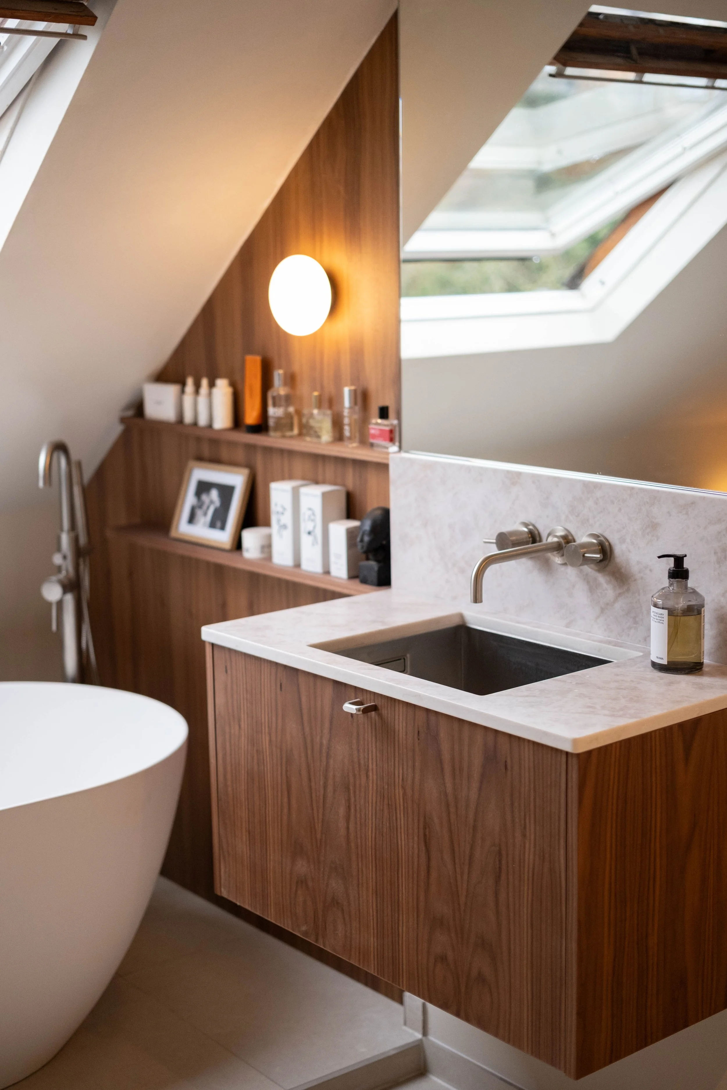 A modern bathroom vanity with a white countertop, a black sink, and a wooden cabinet with a vertical grain. There are various bottles and framed pictures on the shelf above the vanity. A skylight window is visible on the ceiling, allowing natural lig