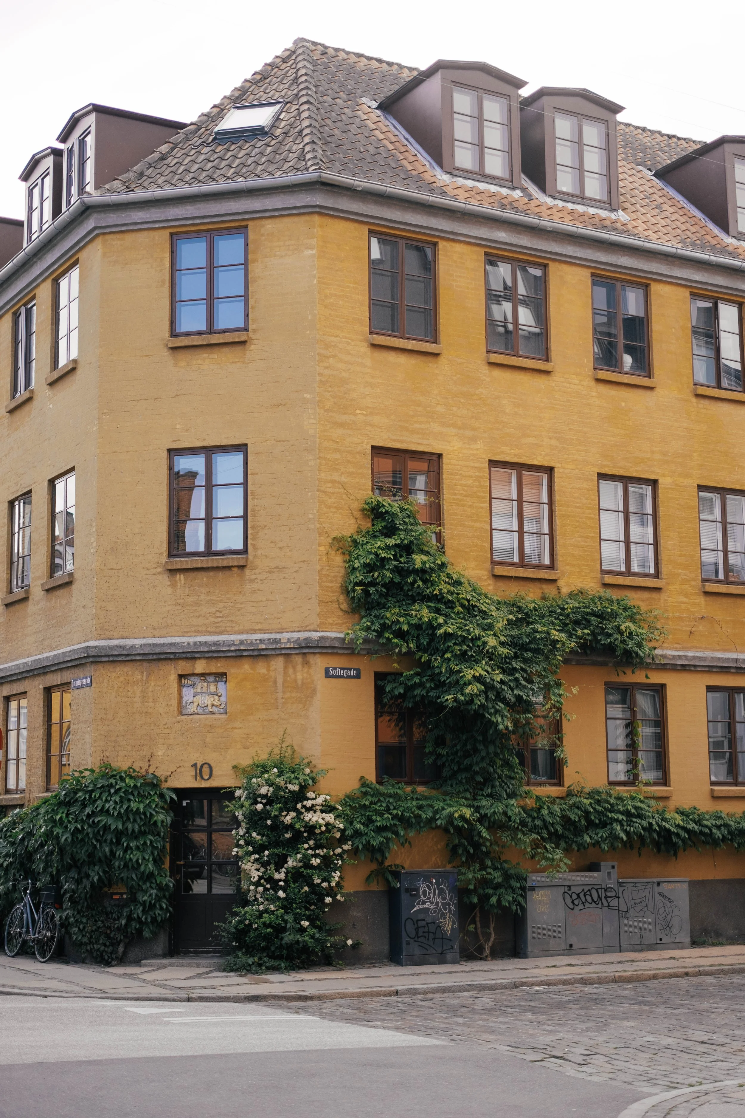 Three-story yellow brick building with multiple windows, some with brown frames and shutters, and a sloped roof with dormer windows. The building is surrounded by green shrubs and trees. There is a street corner at the front with graffiti-covered ele