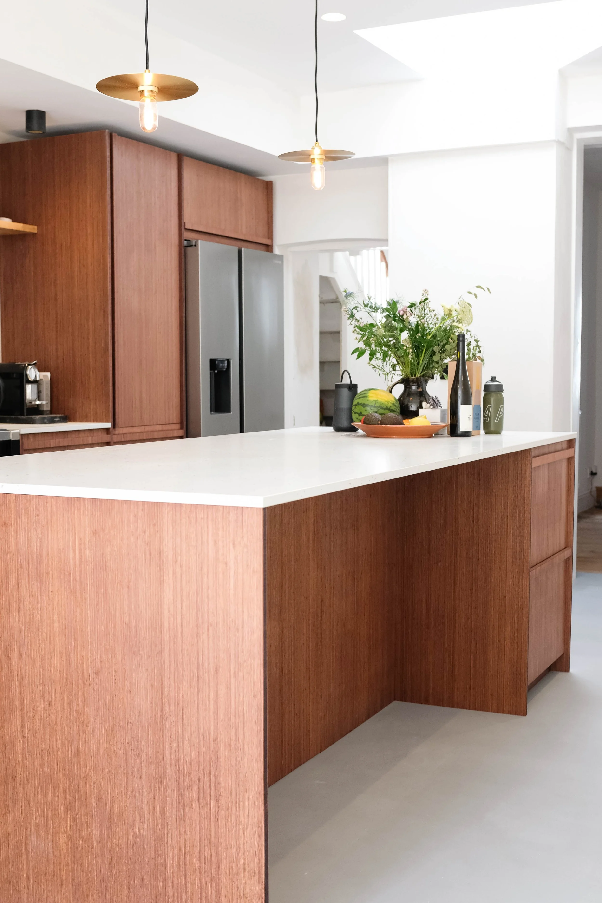 A modern kitchen with wooden cabinets, a white island counter, a stainless steel refrigerator, pendant lights, and a vase of flowers on the counter.