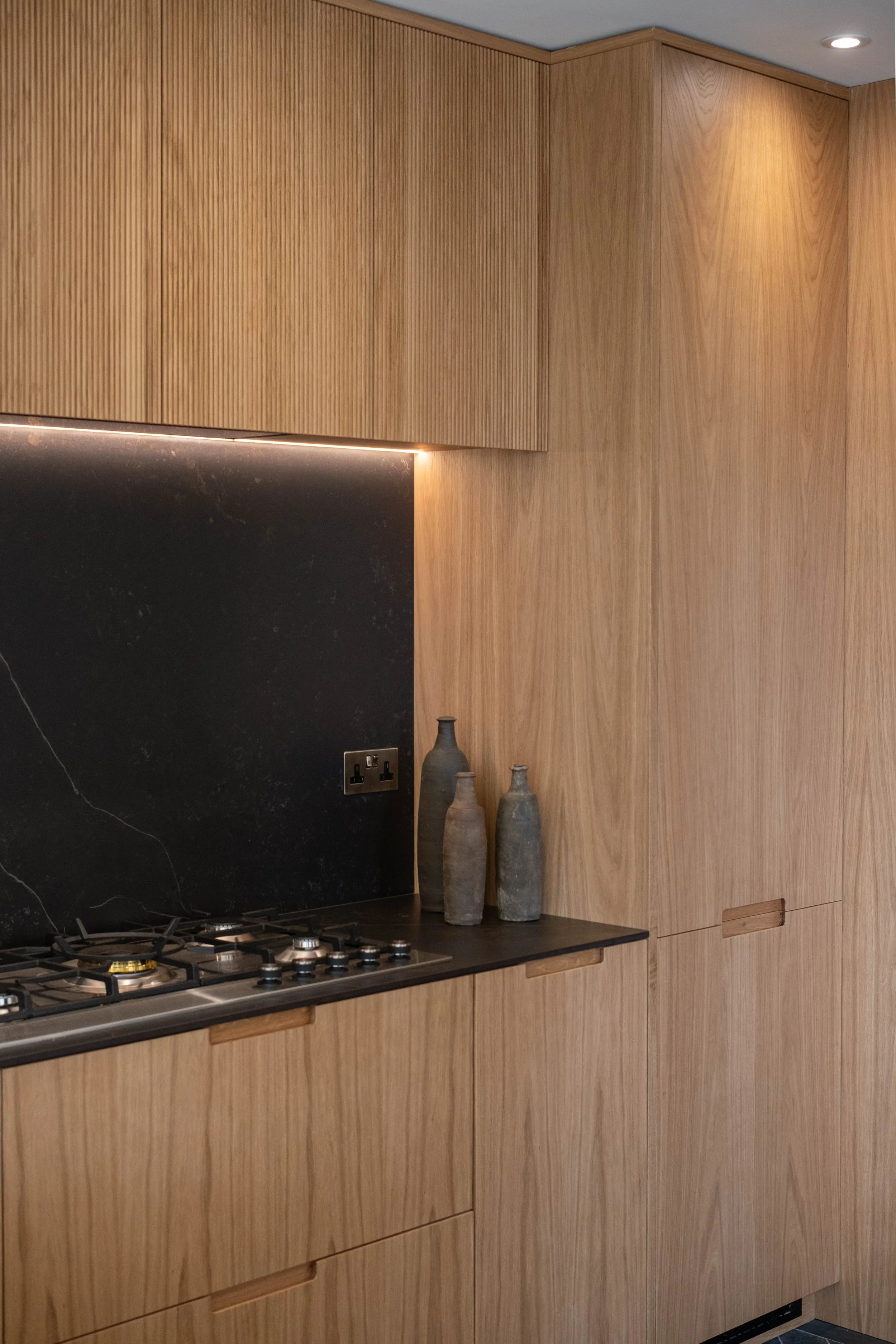 Modern kitchen with wooden cabinets, a black marble backsplash, and three ceramic vases of varying heights on the counter, under a ceiling light.