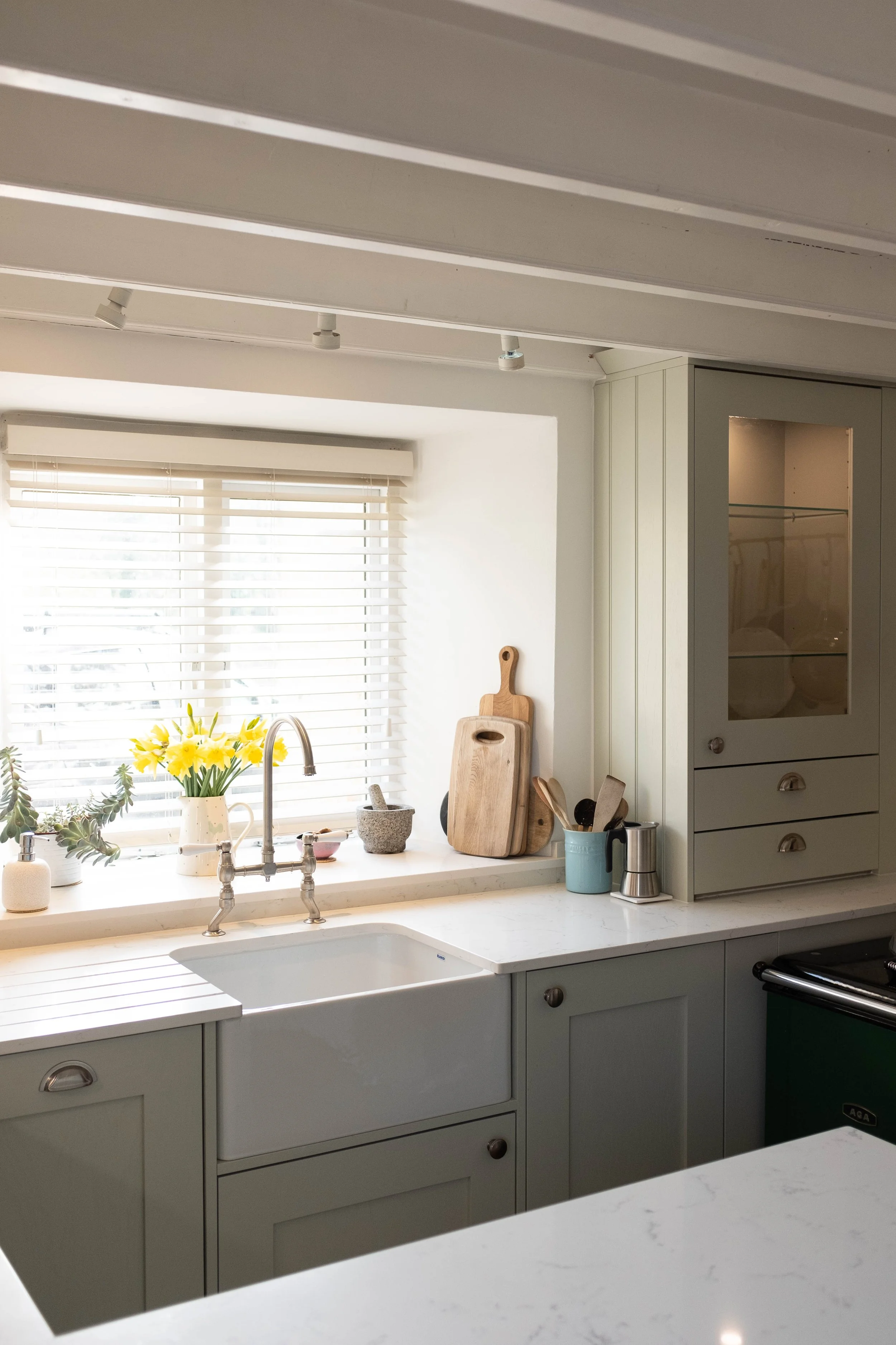A kitchen with a white countertop, light green cabinets, and a window with blinds. On the countertop are a vase of yellow flowers, cutting boards, kitchen utensils, and a small appliance.