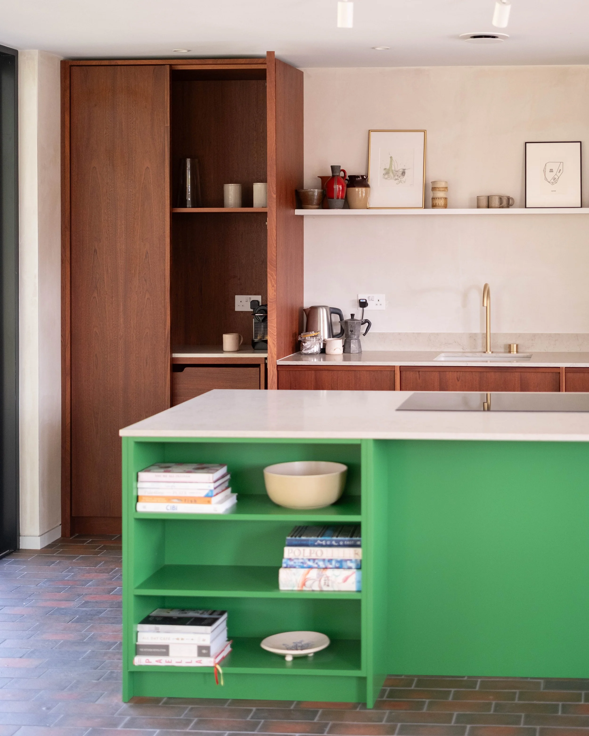 A kitchen with a green island, open shelves with books and dishes, a wooden cabinet with vases and cups, a white countertop with a kettle and moka pot, and framed artwork on the wall.