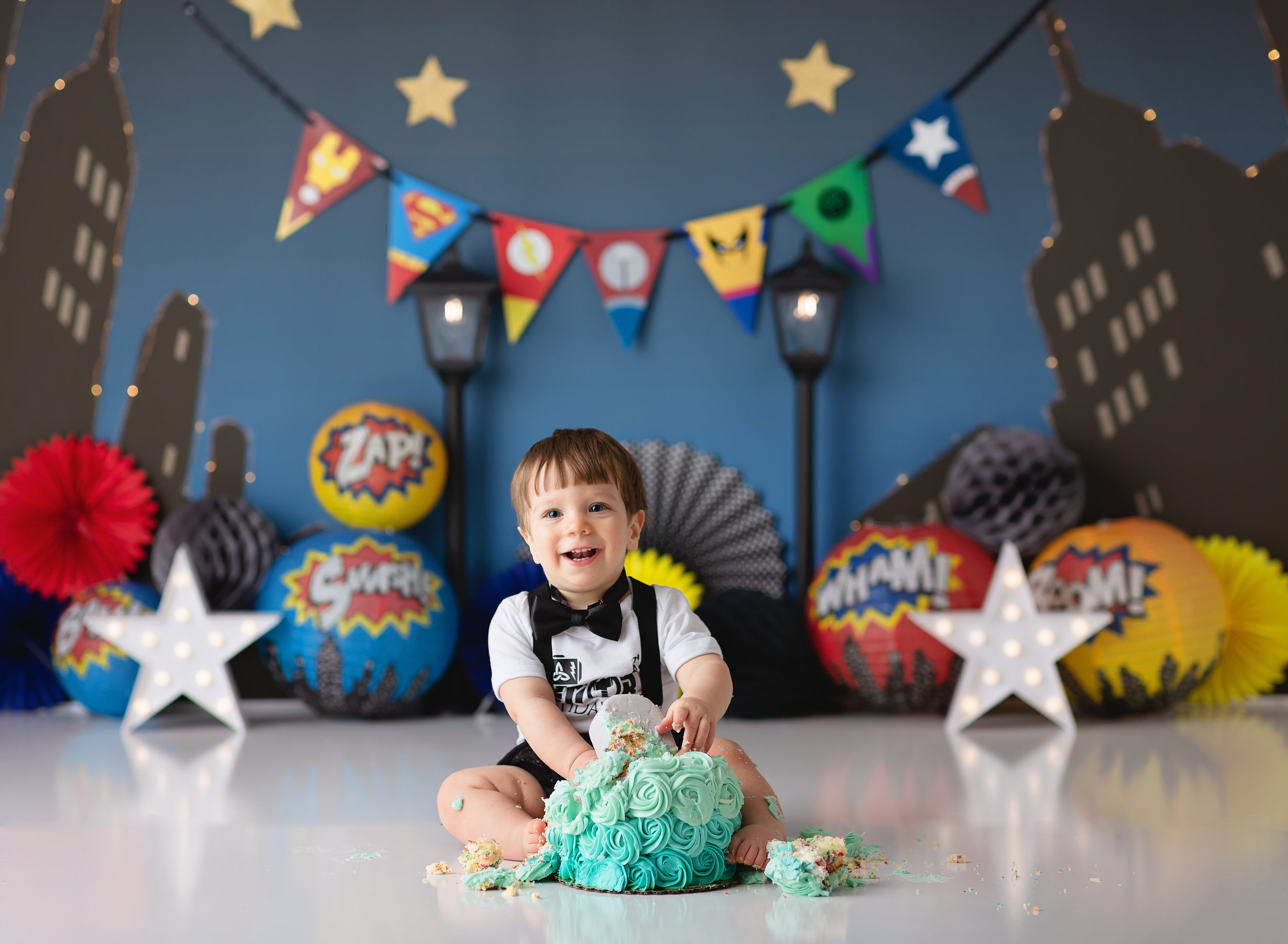  Toddler enjoying his cake with superhero balloons and a cityscape behind him during his superhero cake smash 