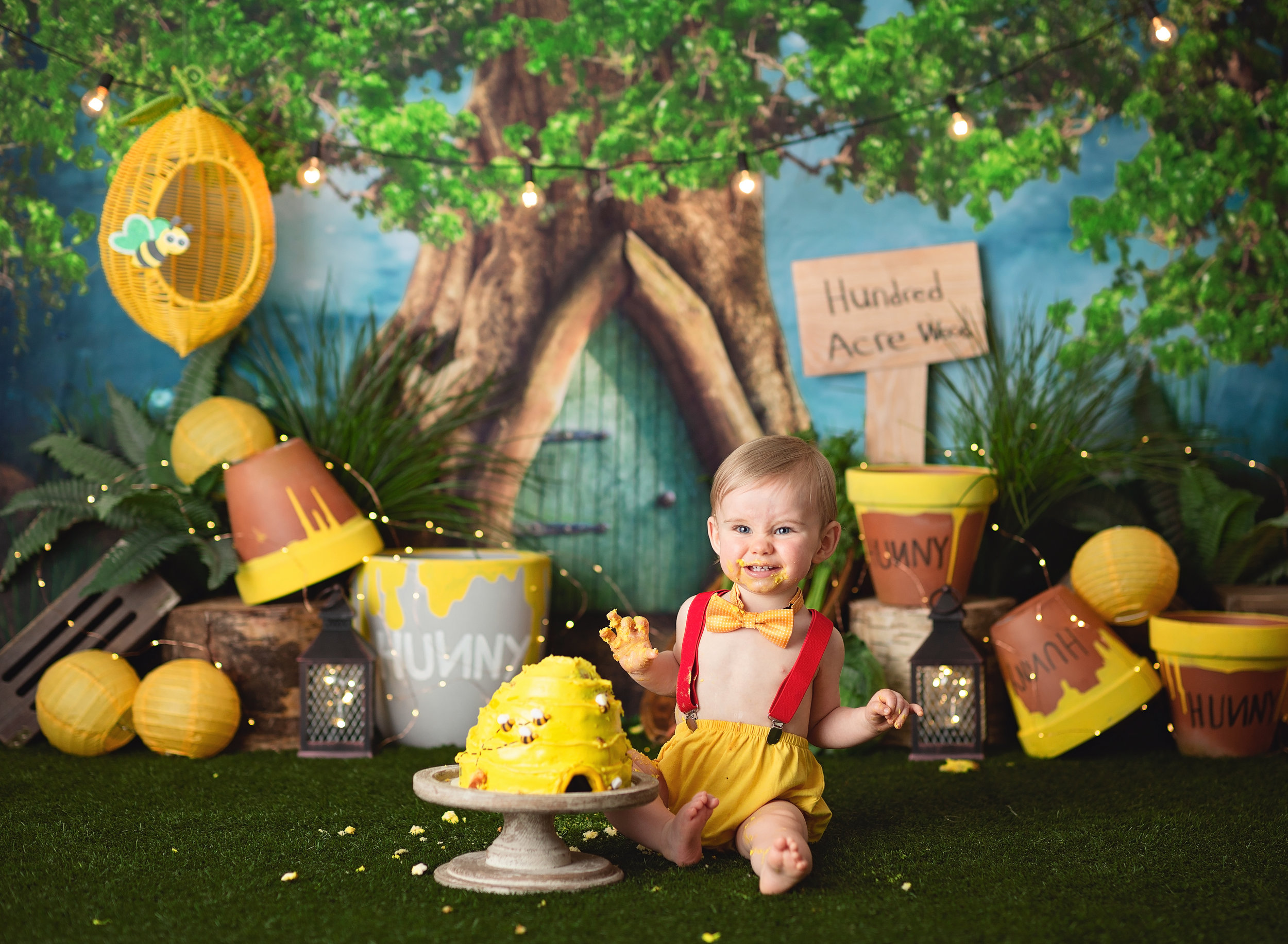  Baby boy dressed like Christopher Robin grins while seated beside a beehive-shaped cake with a Hundred Acre Wood theme in the background 