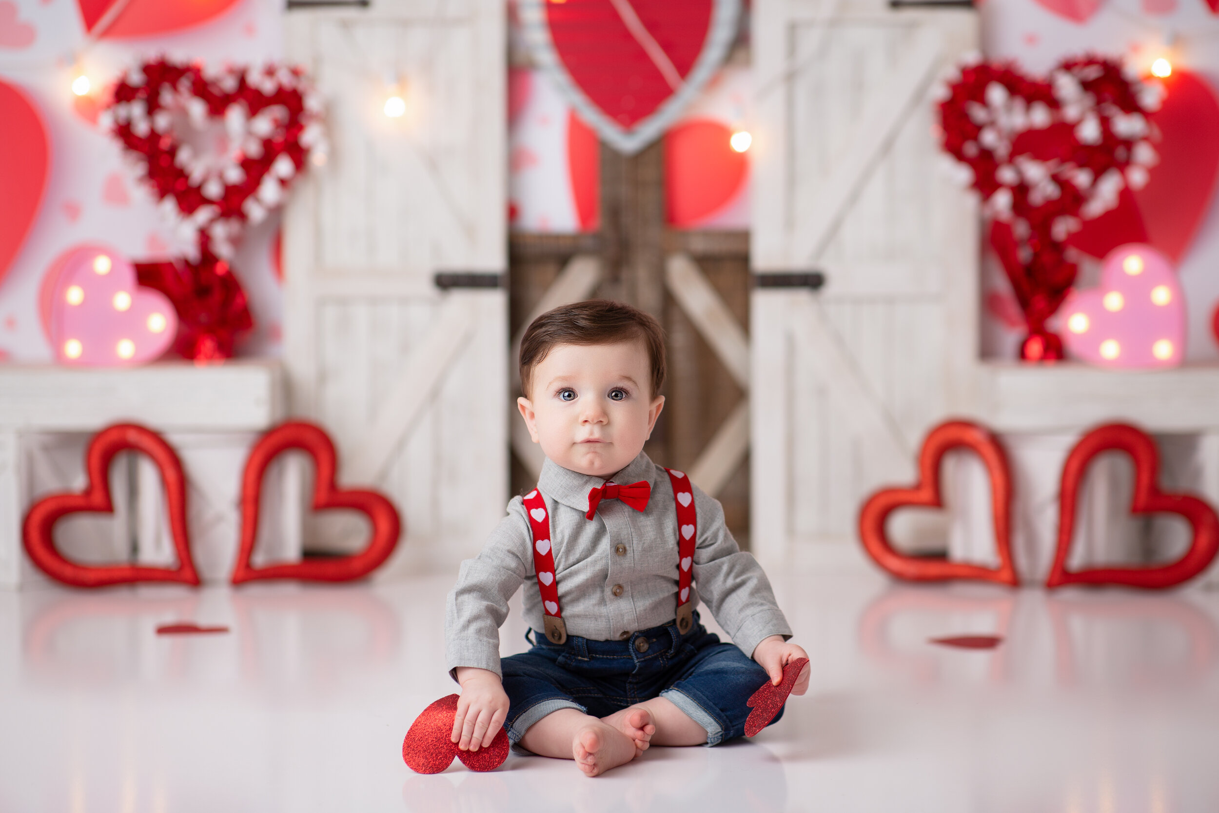  Toddler in red suspenders against a backdrop of hearts during a cake smash shoot near Valentine’s day 