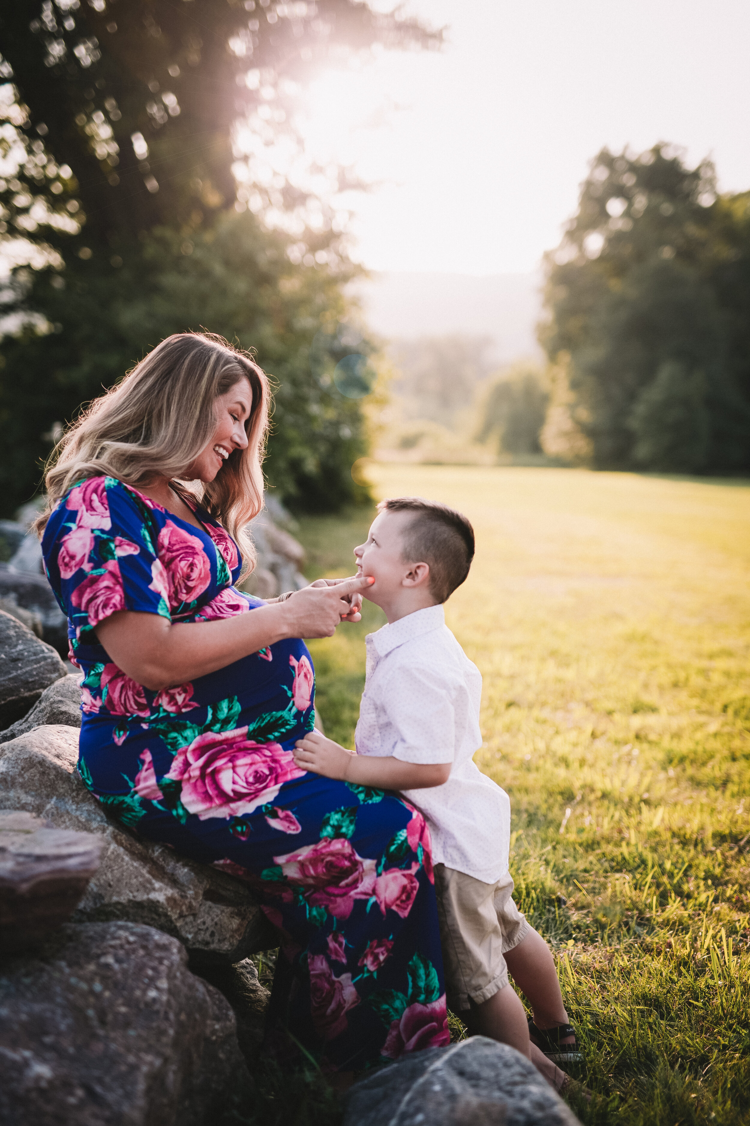  Pregnant mom seated on a rock wall laughs as she looks into her little boy’s face 