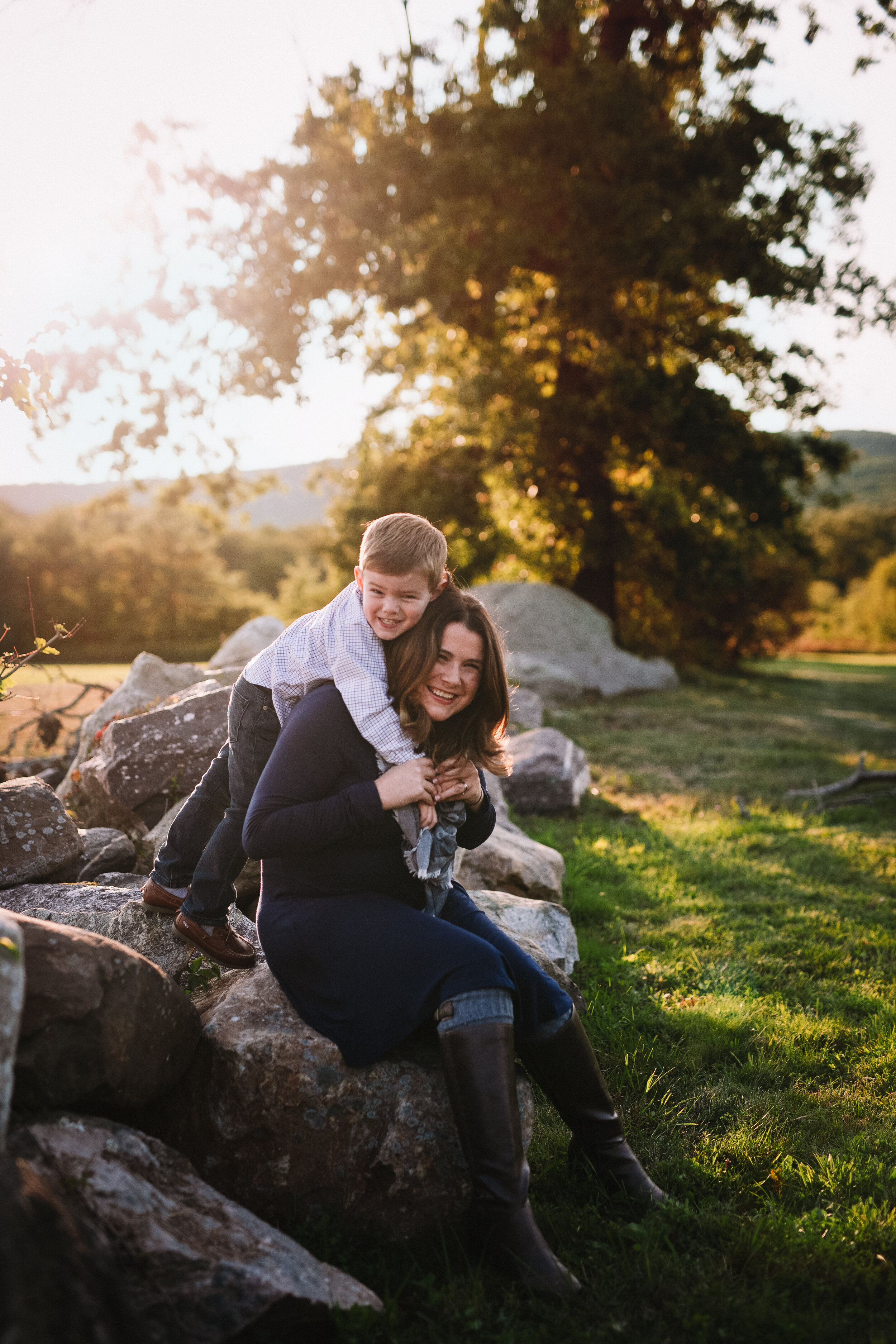  Little boy enthusiastically hugs his mom from behind as she sits on a large boulder in a North Jersey field 