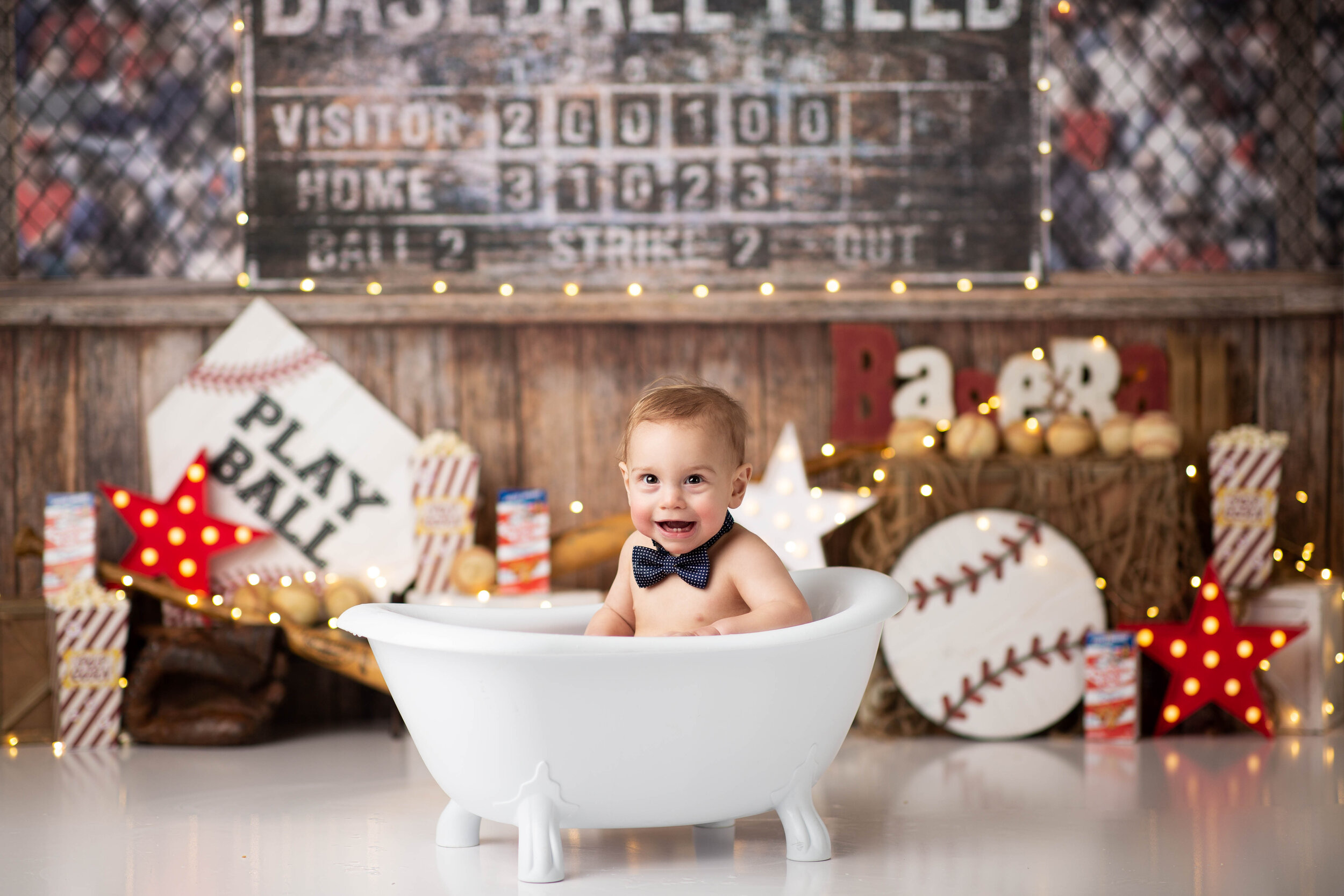  Baby boy splashing in the tub with a scoreboard and baseball paraphernalia in the background 