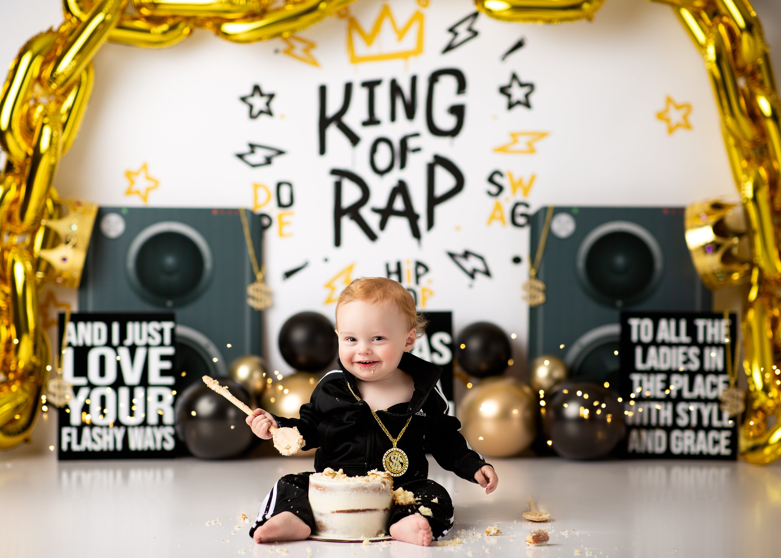  Redhaired baby boy dressed in black and waving a wooden spoon during his King of Rap cake smash 