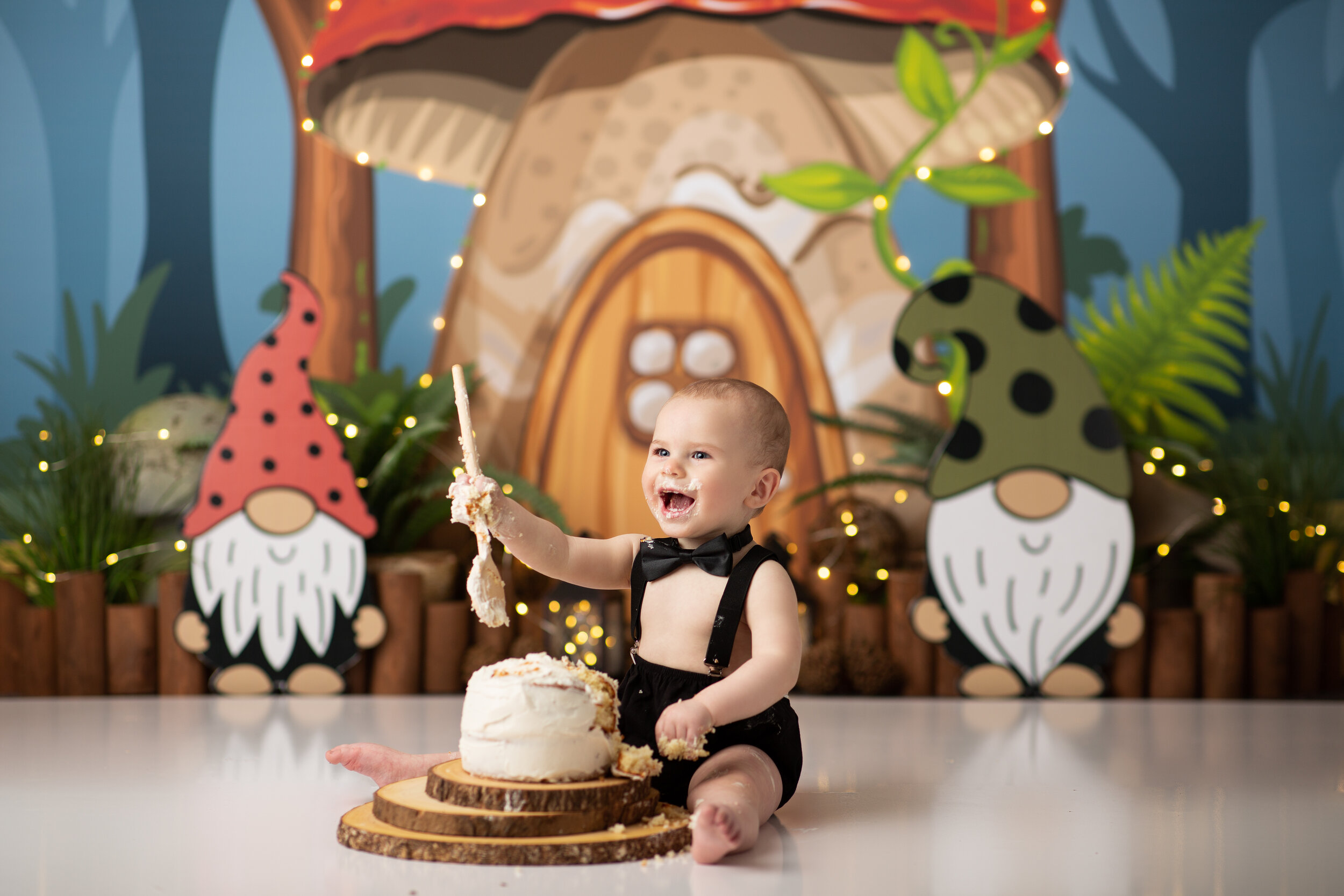  Baby laughing and holding wooden spoon while he eats his first birthday cake against a backdrop of mushrooms and gnomes 