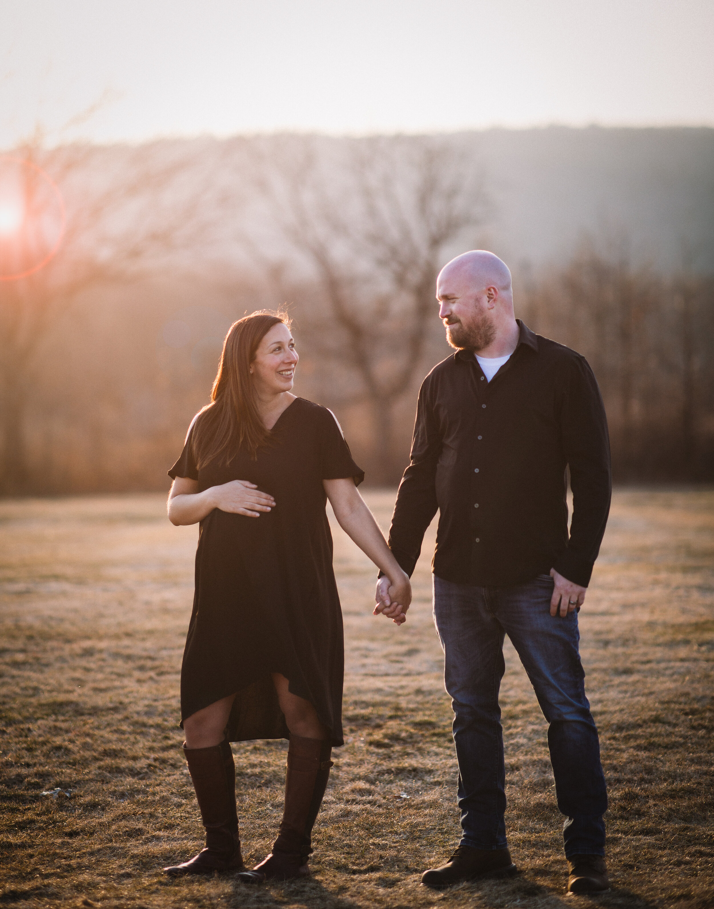  Husband and wife in casual attire holding hands in a sunny field during a maternity photo session 