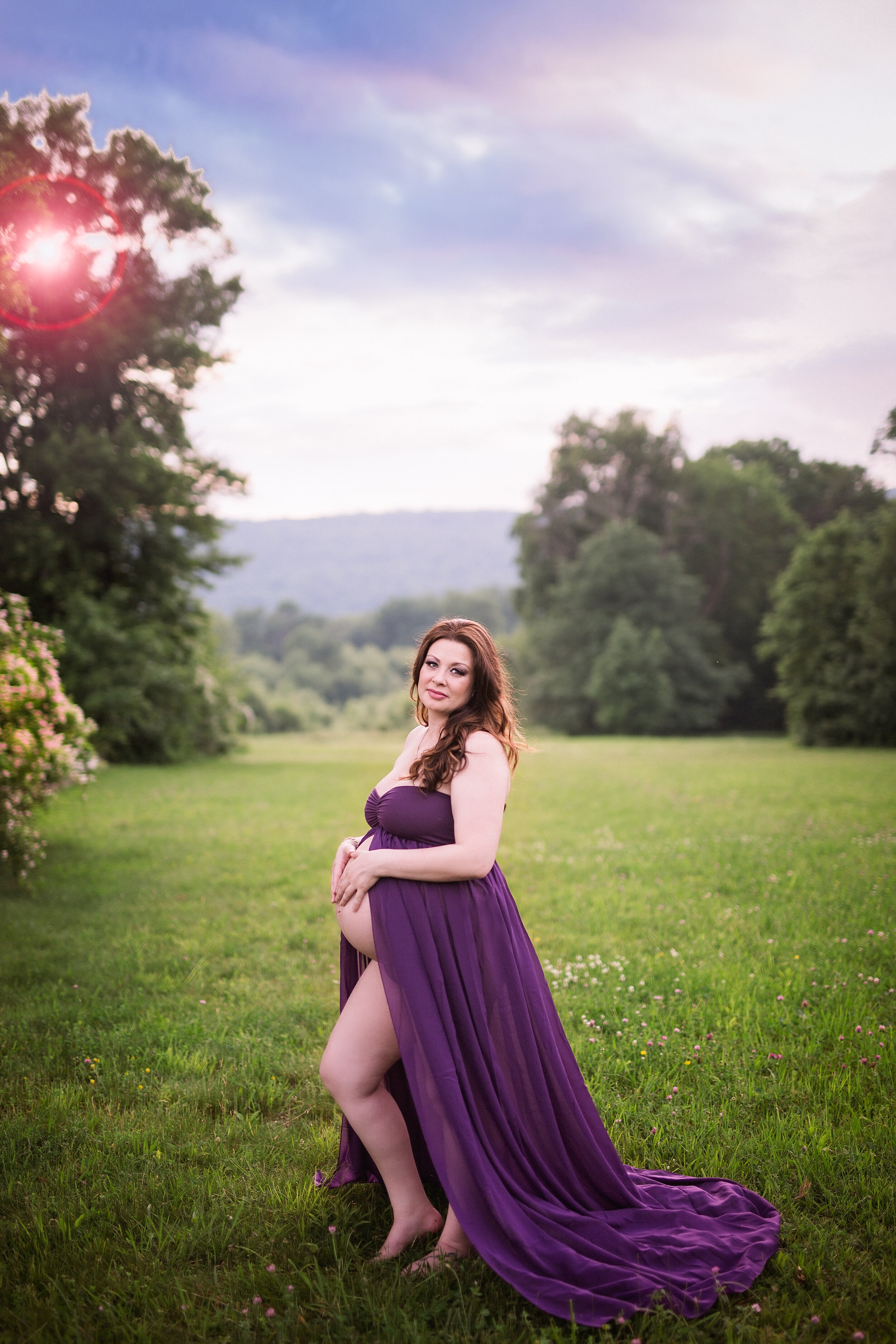  Woman standing in open field with trees and fields behind her as she poses in a flowing purple maternity dress 