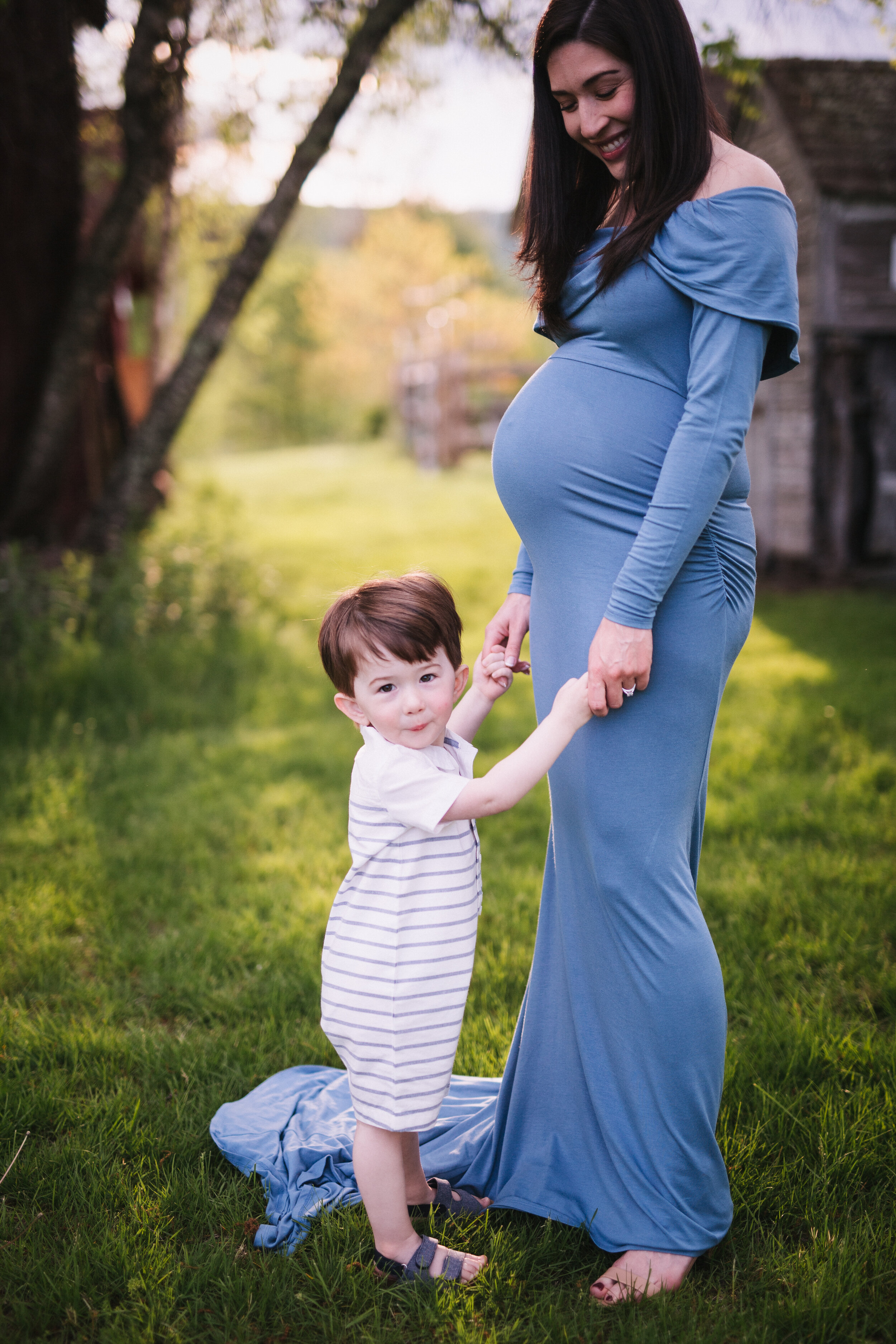  Mother in flowing sky-blue maternity dress holds her little boy’s hands in a wooded meadow 