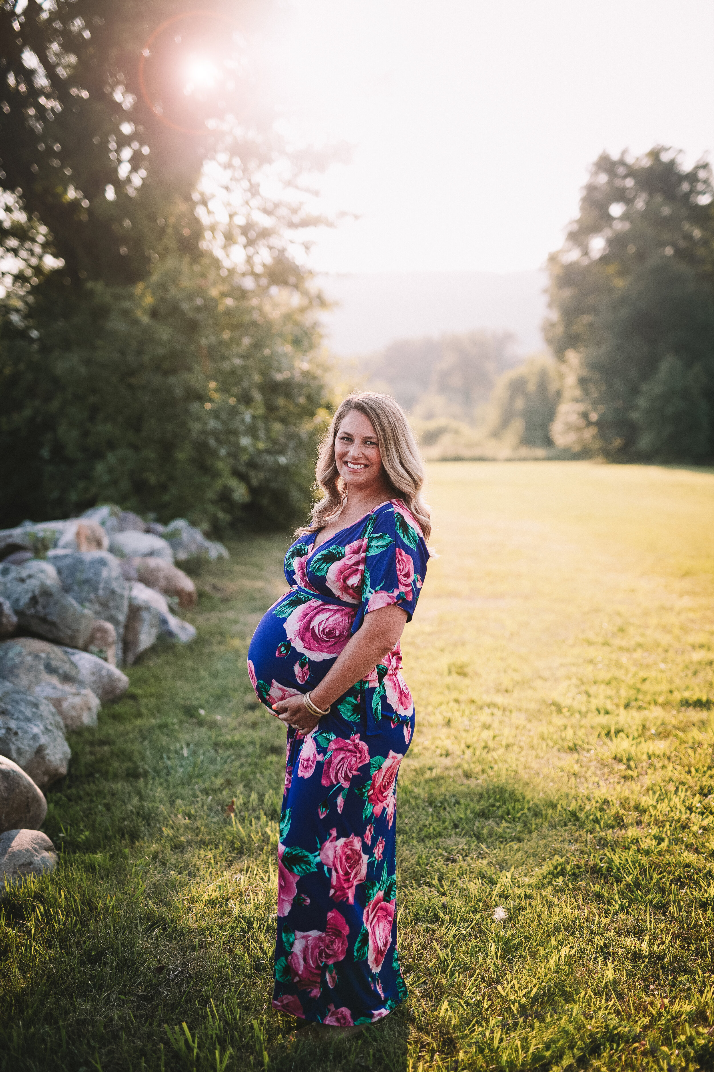  Blonde woman in blue and pink floral dress smiles for photographer during New Jersey maternity photoshoot 
