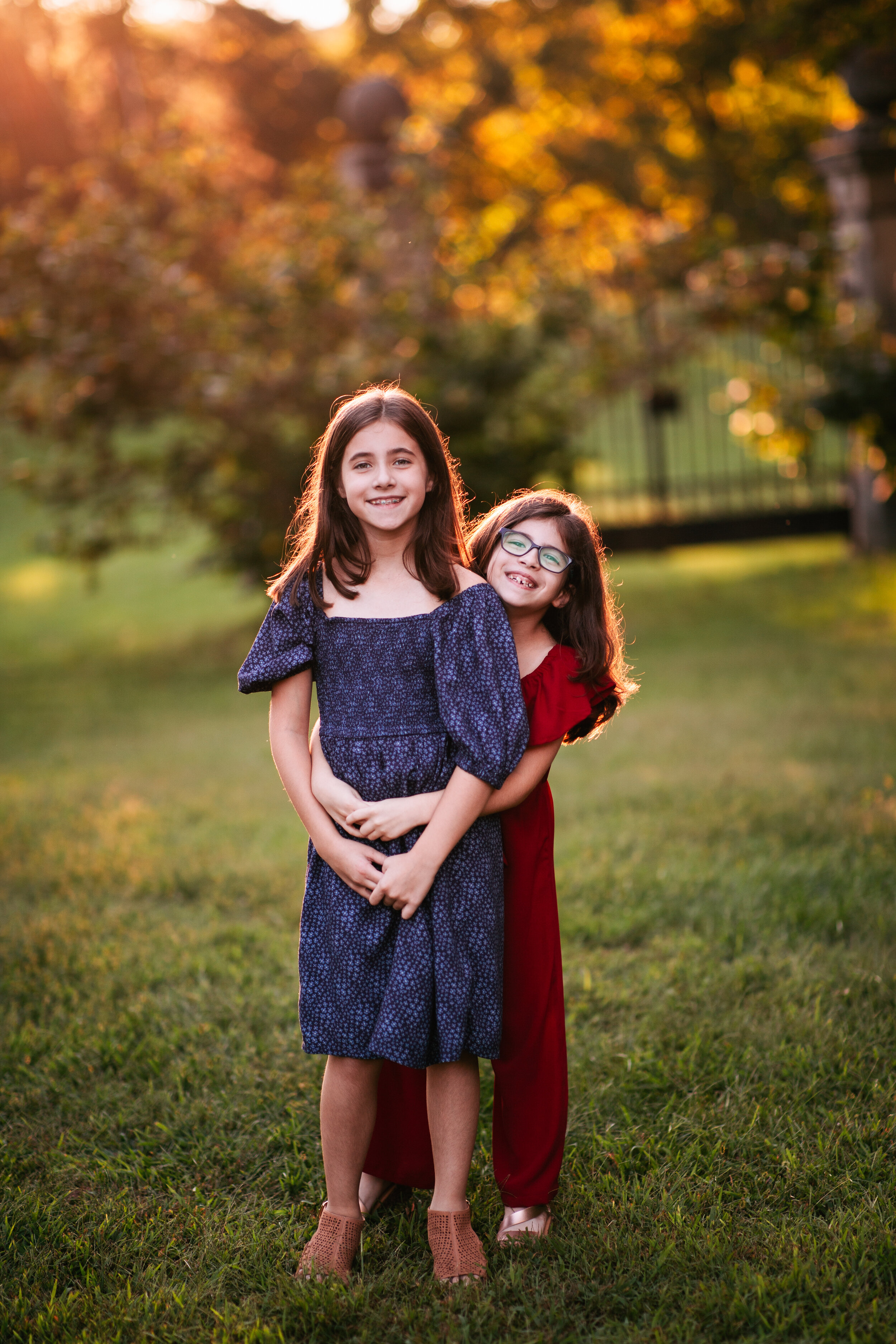  Little girl stands smiling while her young sister wraps her arms around her in a hug from behind 
