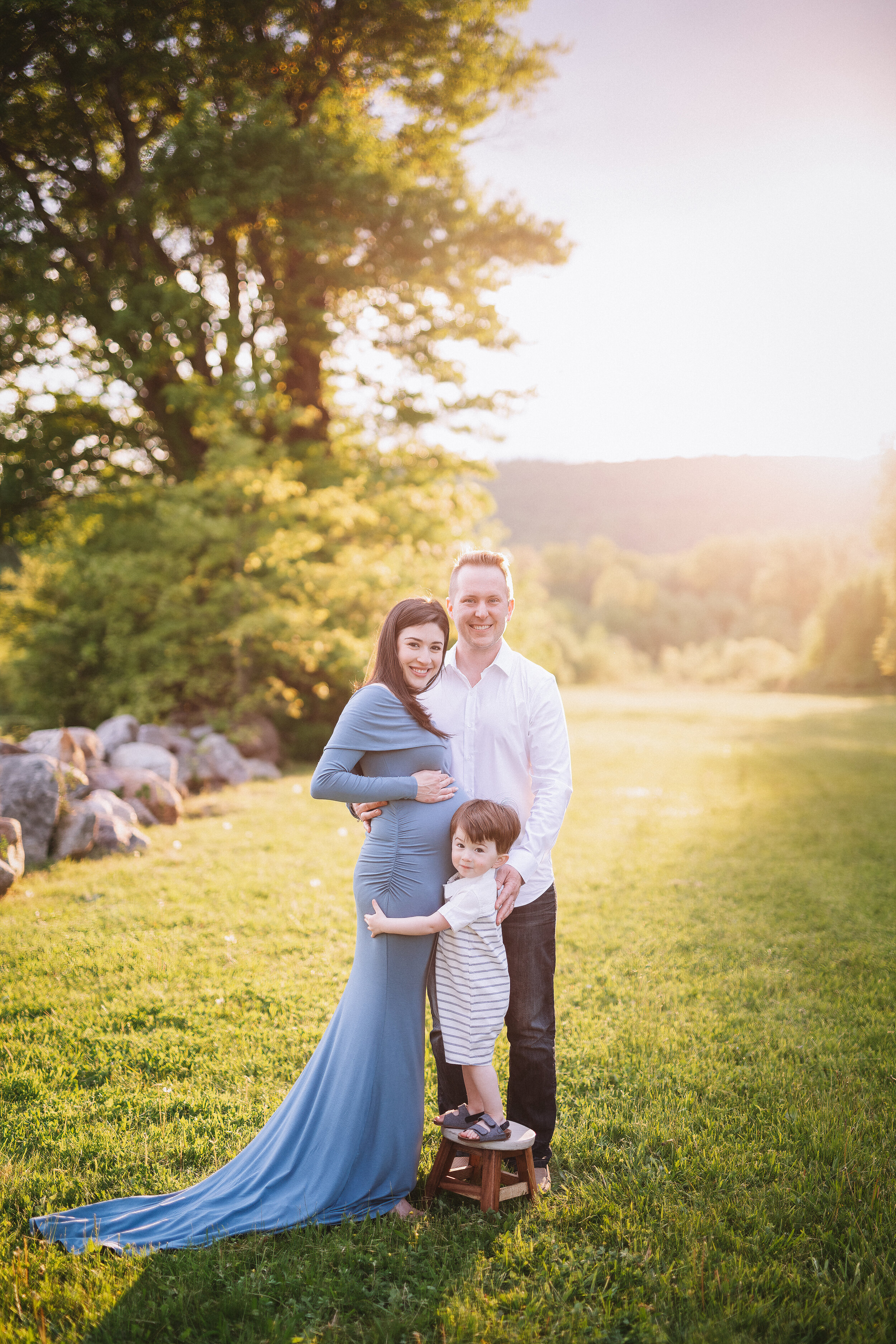  Pregnant mom in flowing sky blue dress stands with her husband and young son in a beautiful sun-soaked field 