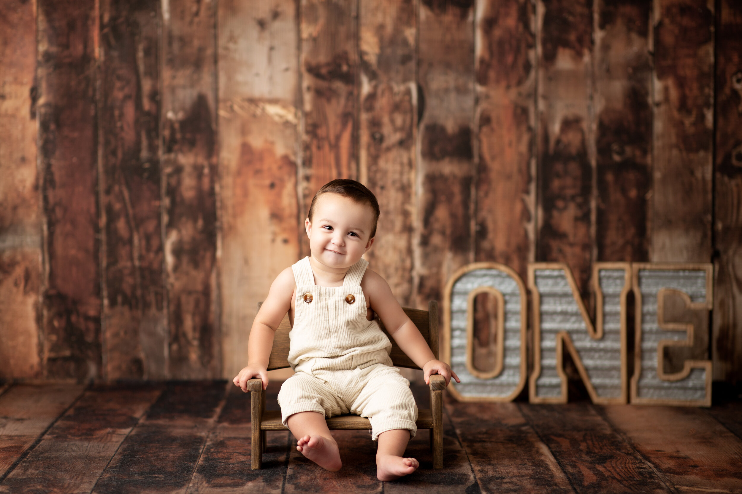  Little boy seated against rustic wood backdrop with a metal sign spelling out the word “one” 