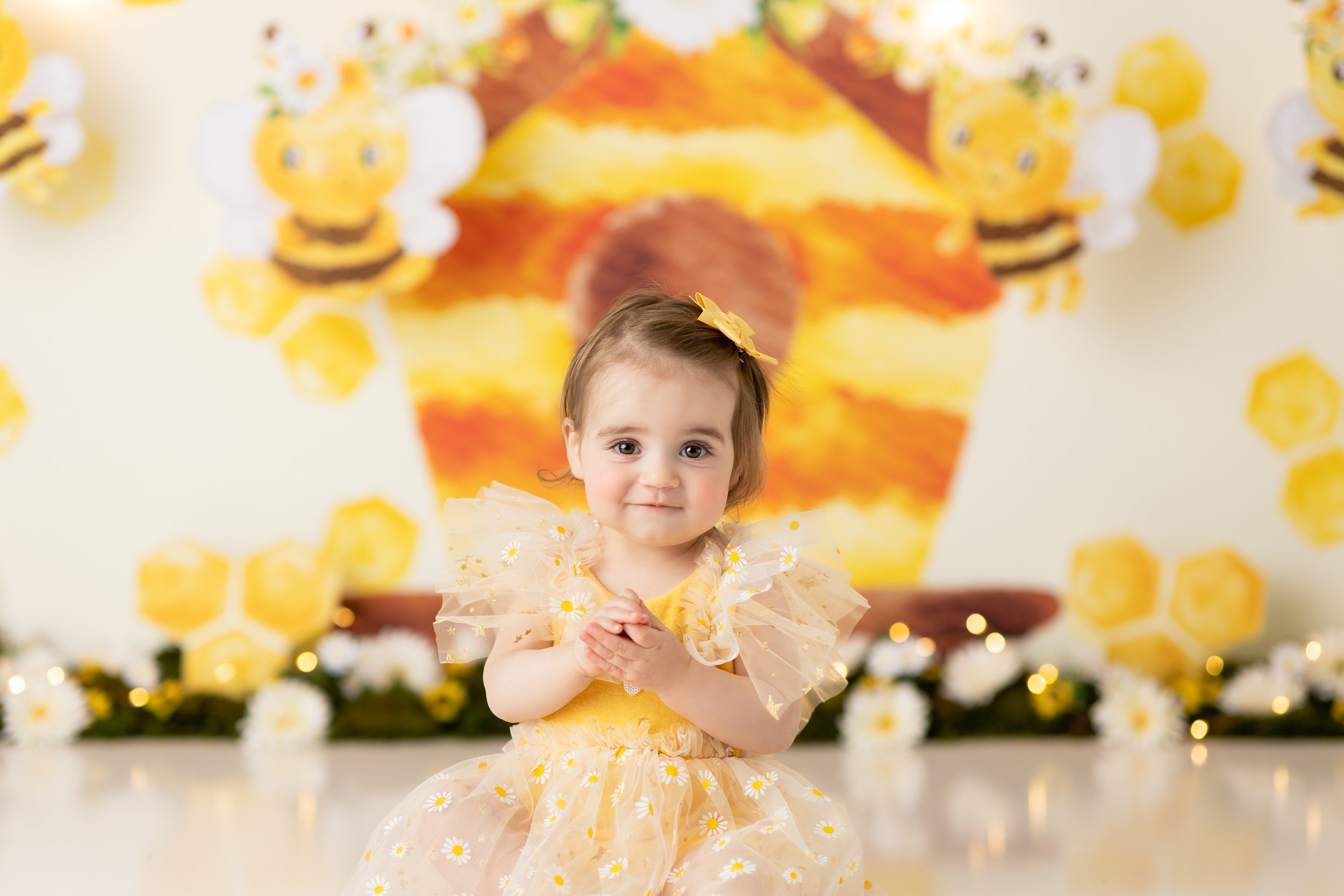  Smiling baby girl all dressed in yellow clasps her hands in excitement during her bumblebee themed custom birthday photo session in the Tri-State area 
