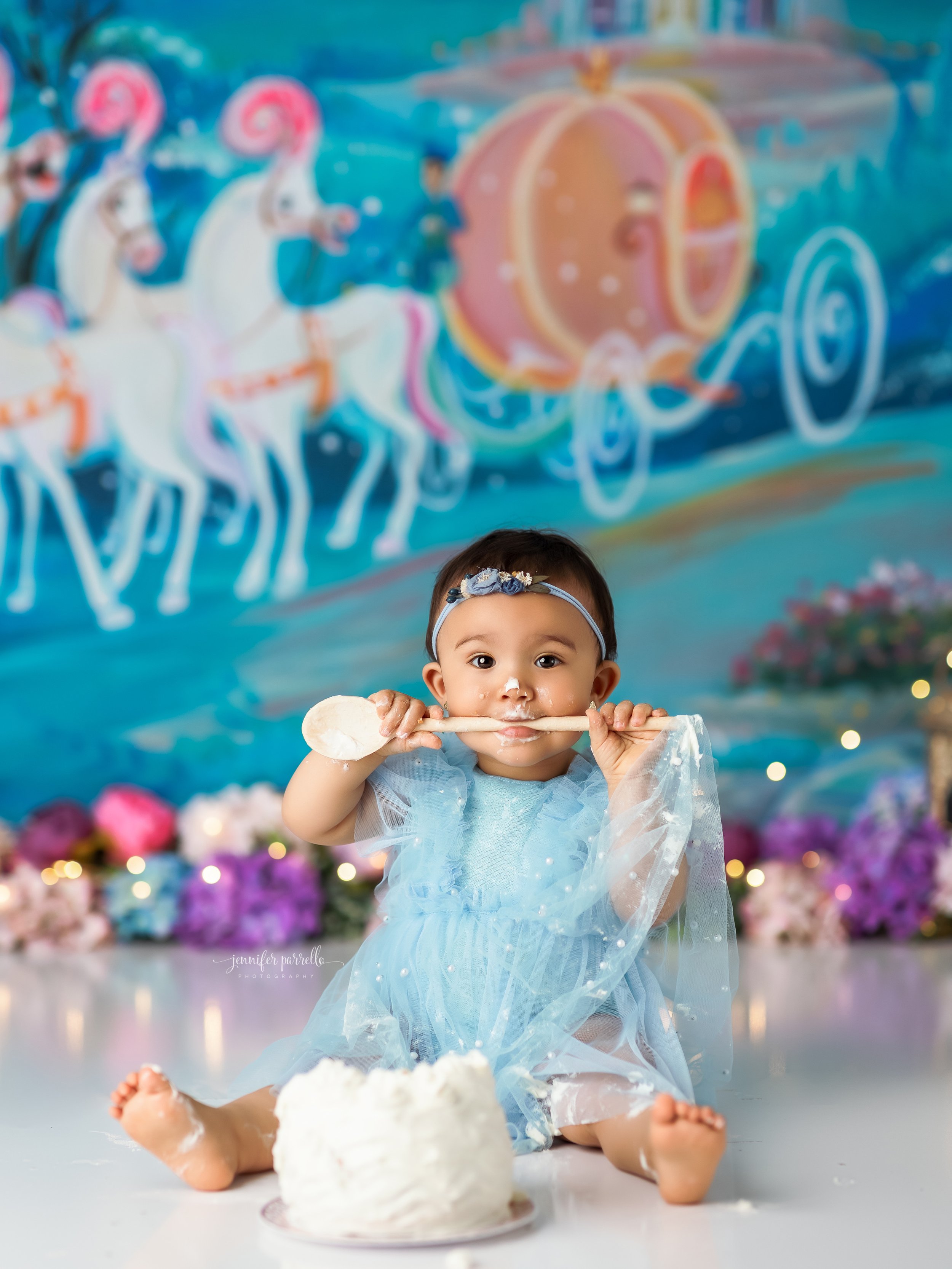  Baby girl in blue dress gnawing on a wooden spoon during her cake smash, with a Cinderella pumpkin carriage on the wall behind her 