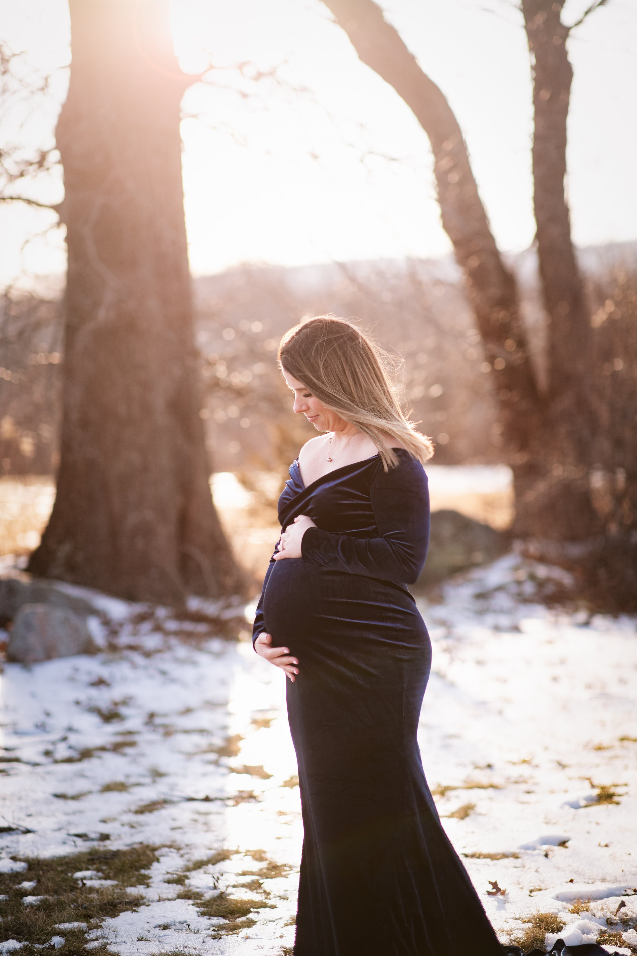  Woman in romantic black maternity gown framed between trees while standing in a snowy field near sunset 