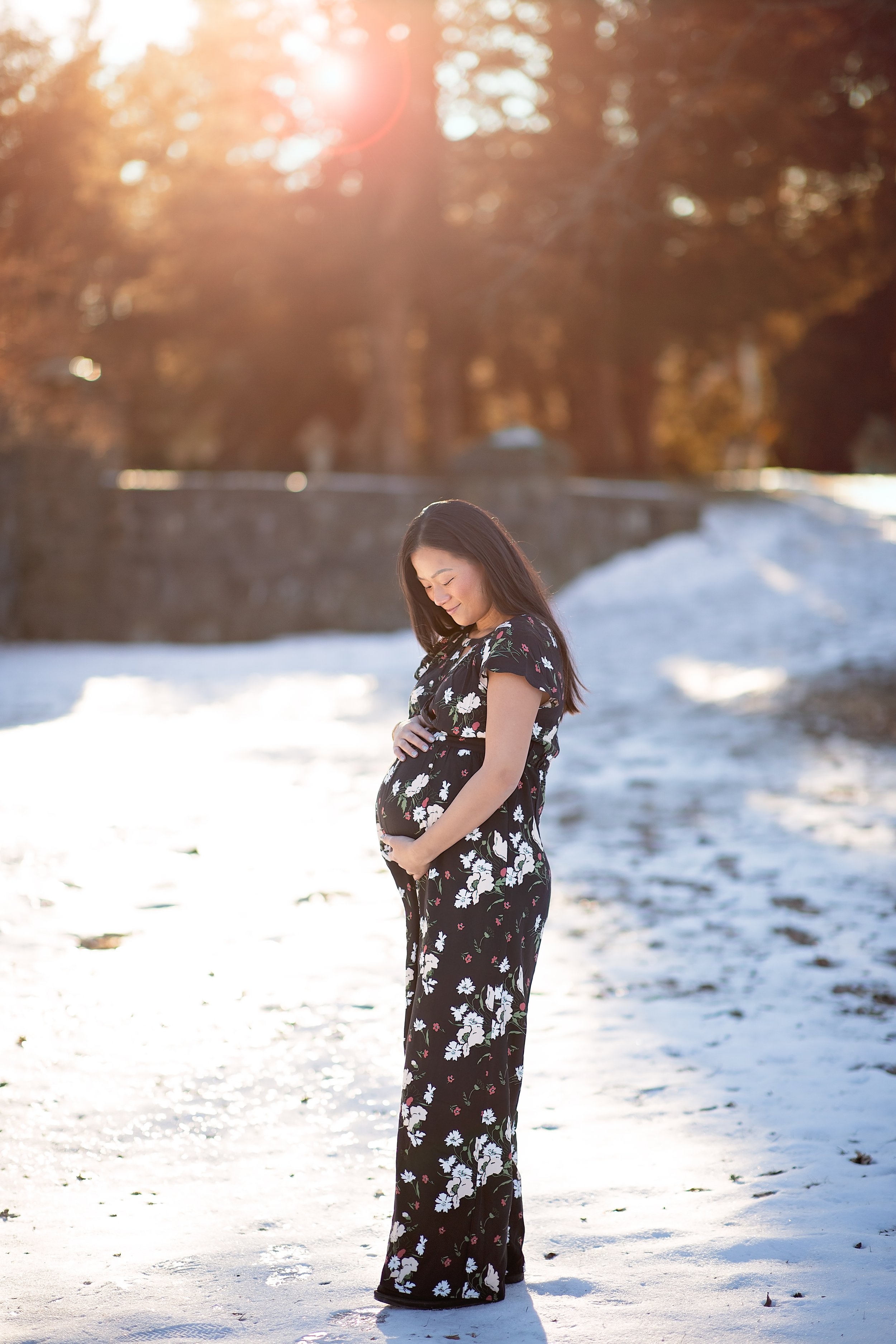  Asian woman in floral maternity maxi dress cradles her belly while standing in a snowy field 