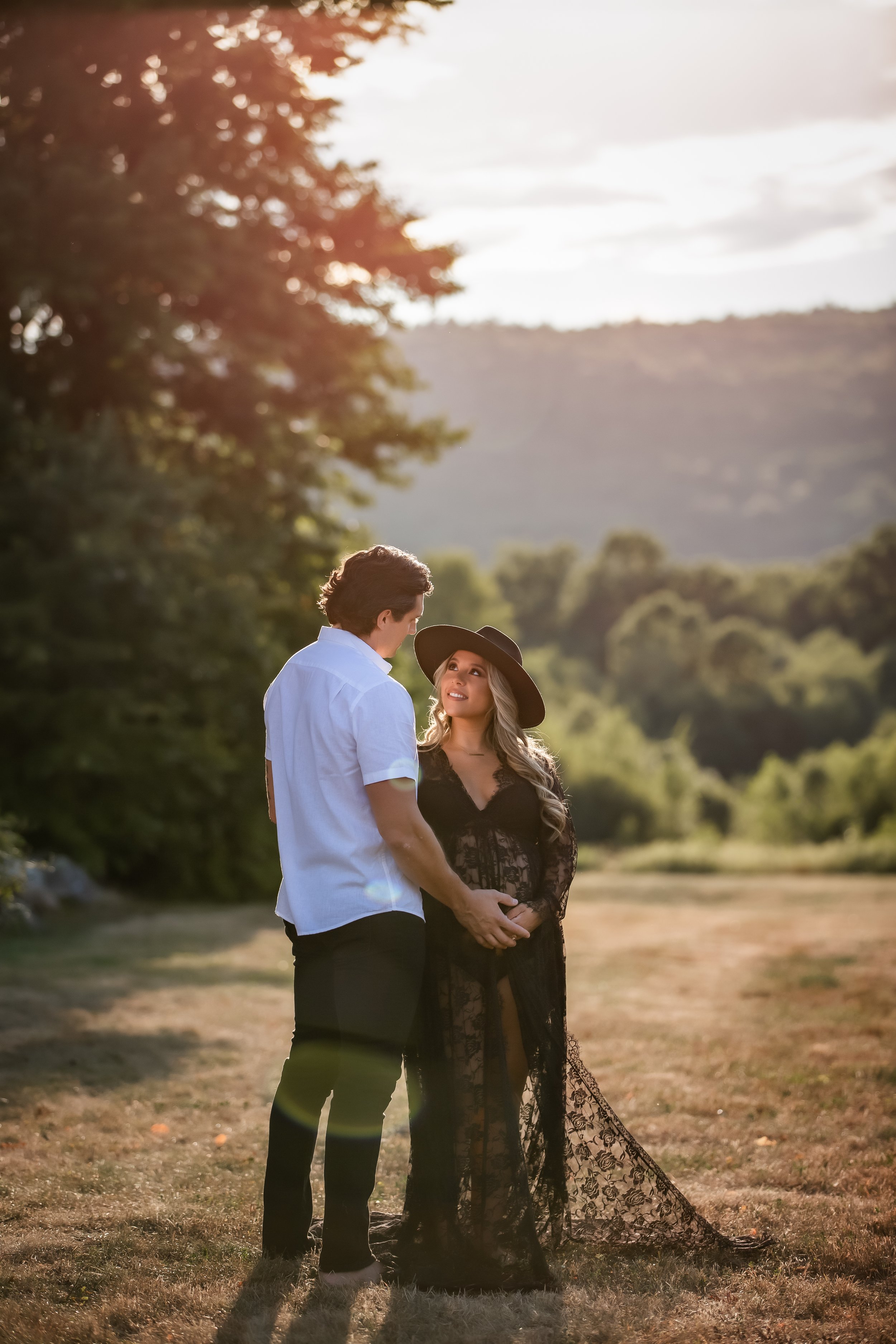  Woman in black maternity gown and cowboy hat gazes up at husband with sunlit hills in the background 