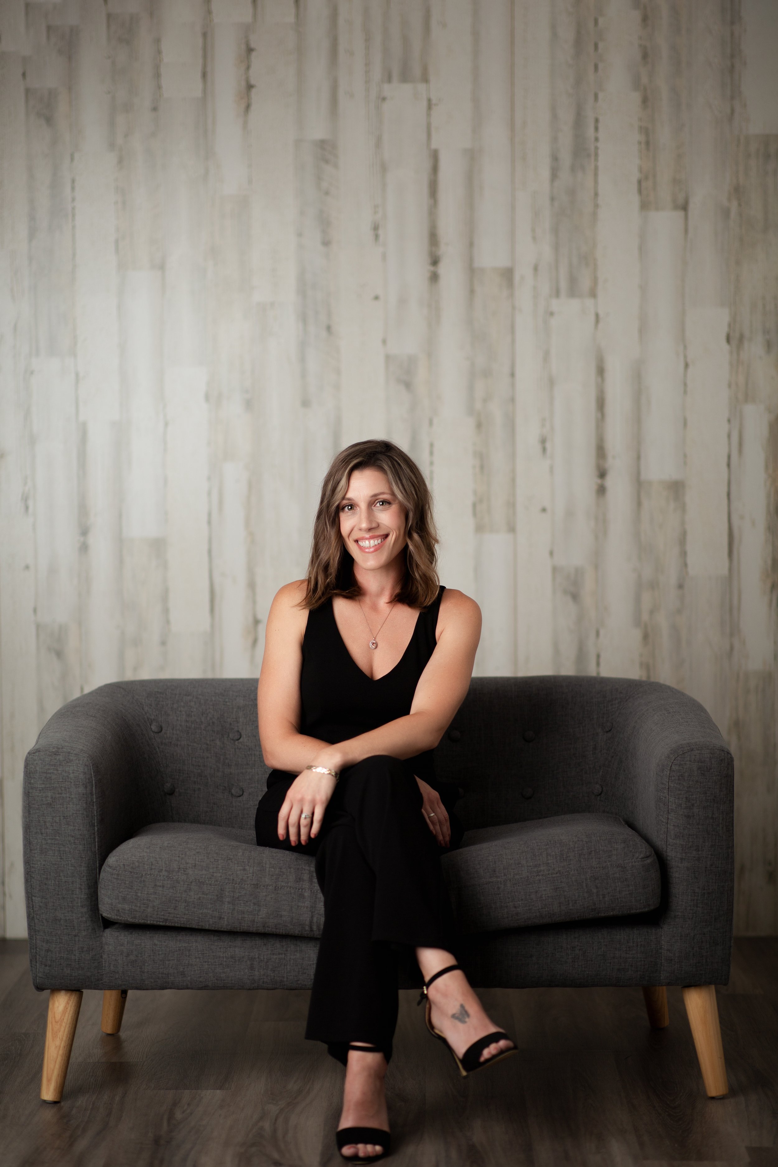  Woman in black pantsuit and heels sitting cross legged on a gray couch 