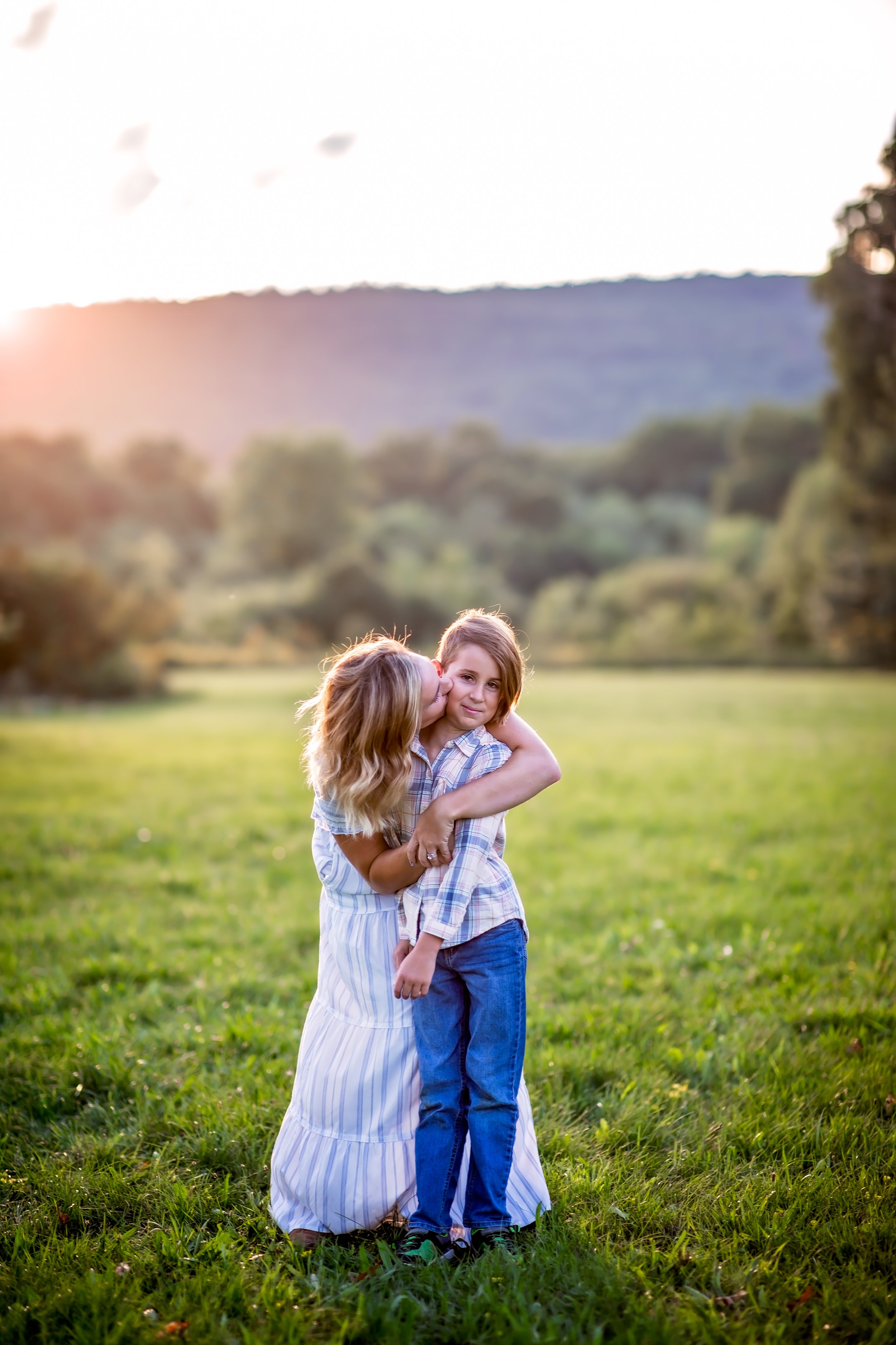  Mom in white dress wraps her arms around her young son and kisses his cheek in a pretty New Jersey field 